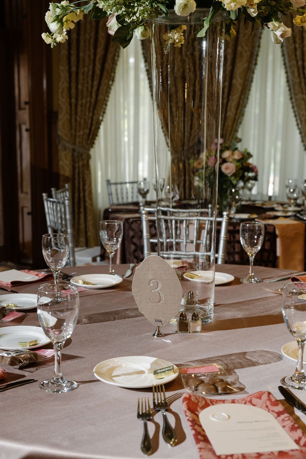 Wedding reception table setup with a tall glass vase filled with flowers as a centerpiece, surrounded by wine glasses, plates, silverware, and a table number card with the number 3.