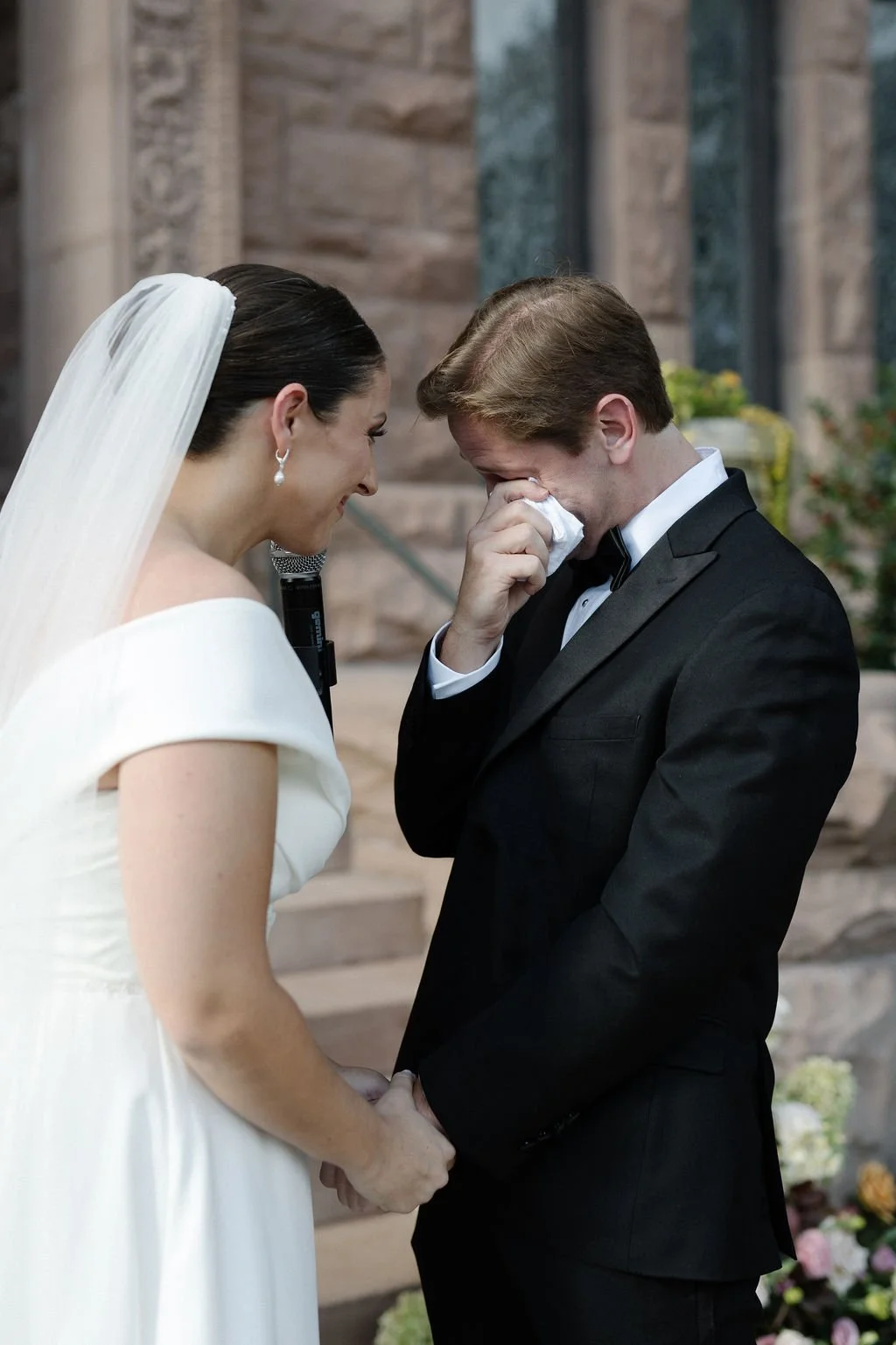 A bride and groom holding hands during their wedding ceremony. The groom is emotional, wiping tears with a tissue, while the bride smiles and looks at him. The bride wears a white dress and veil, and the groom wears a black tuxedo. They are outside, 