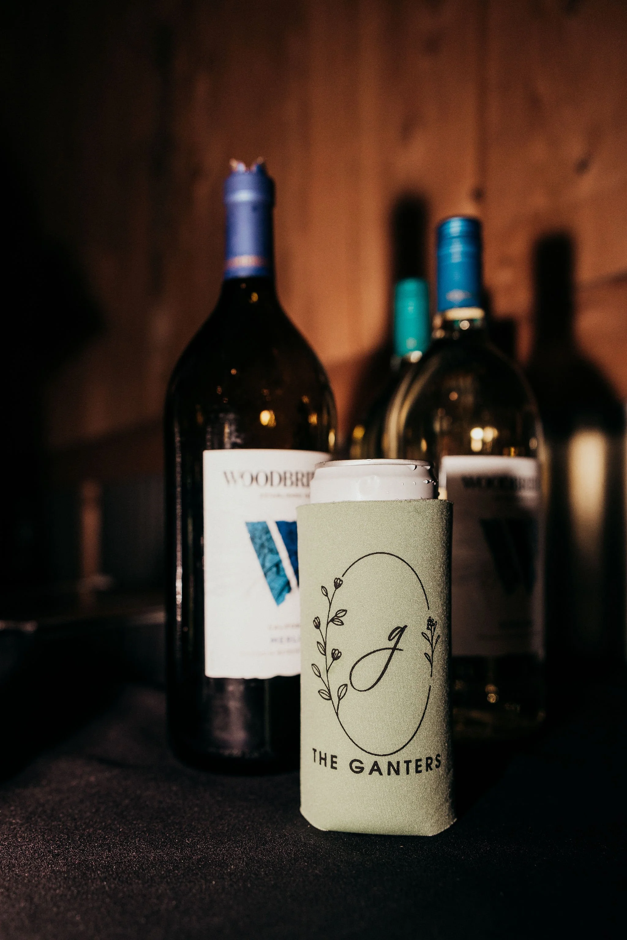 Three bottles of alcohol on a dark surface with one in the foreground wrapped in a white label with decorative black text and design, and two in the background with dark and teal caps. The background is a wooden wall.