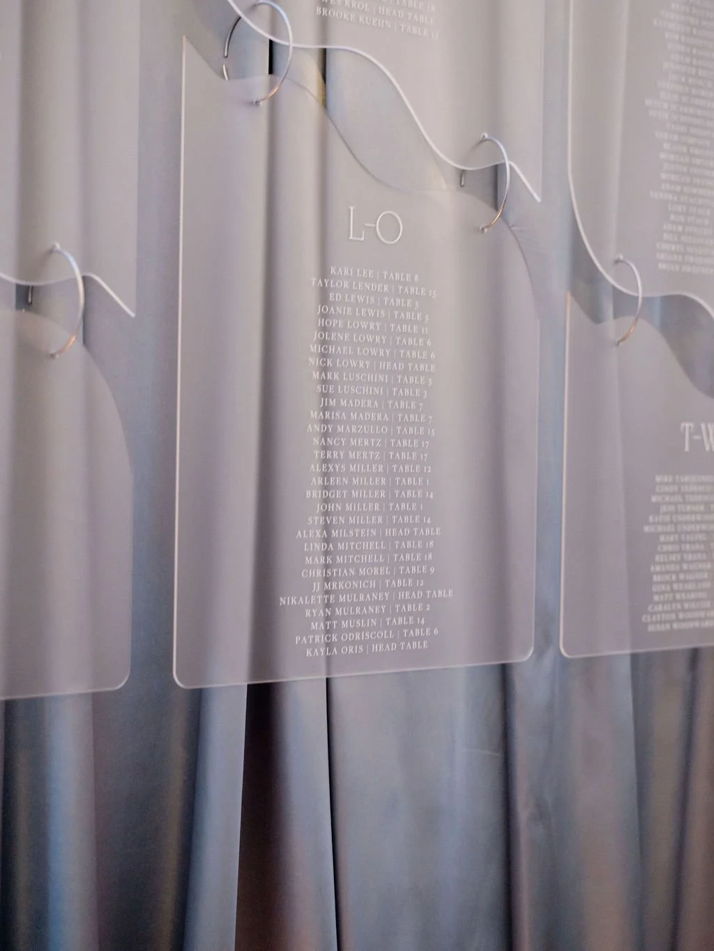 A transparent seating chart with guest names and table numbers, hung on a metallic wall, with decorative white string lights around it.