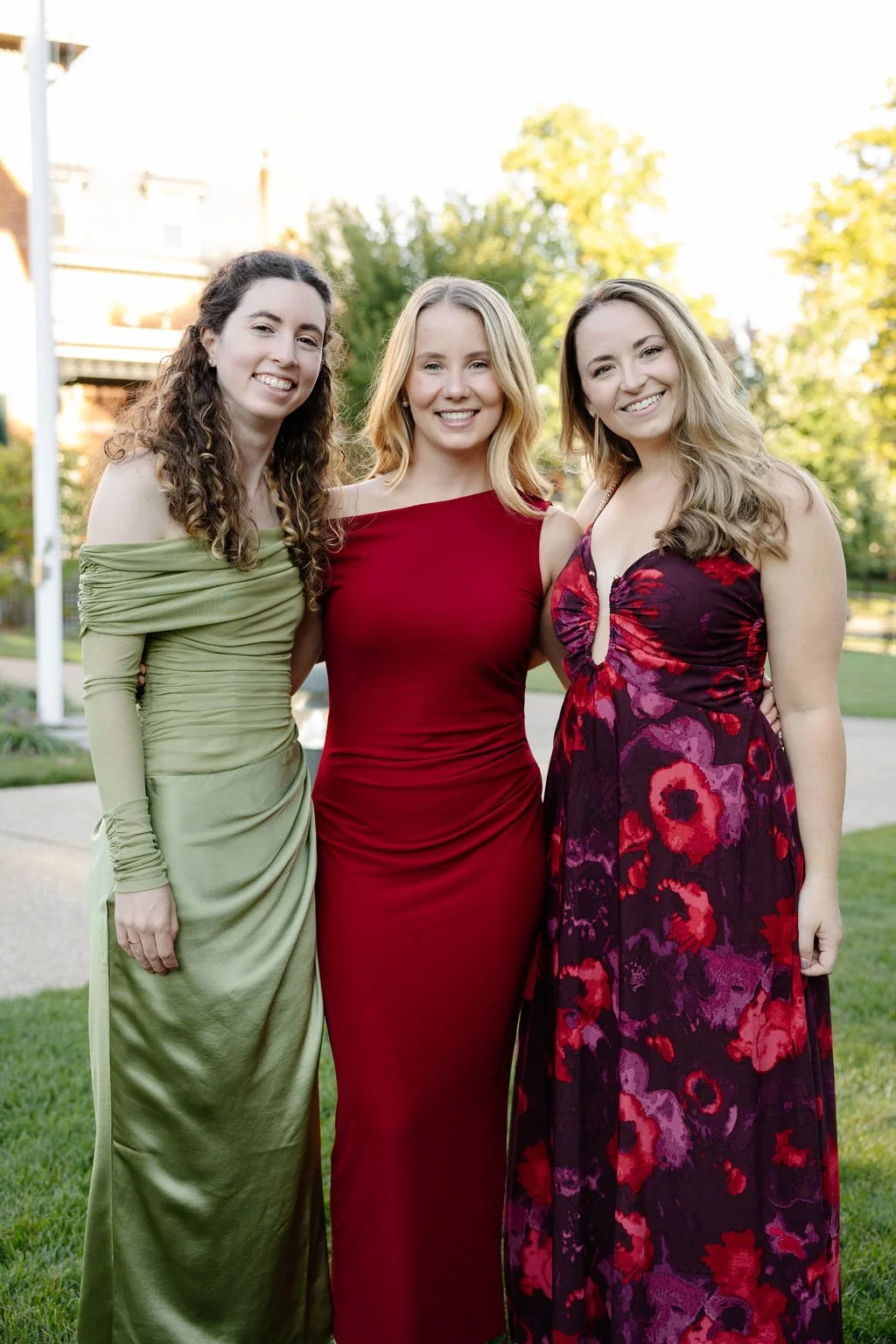 Three women in formal dresses smiling outdoors with trees and houses in the background.