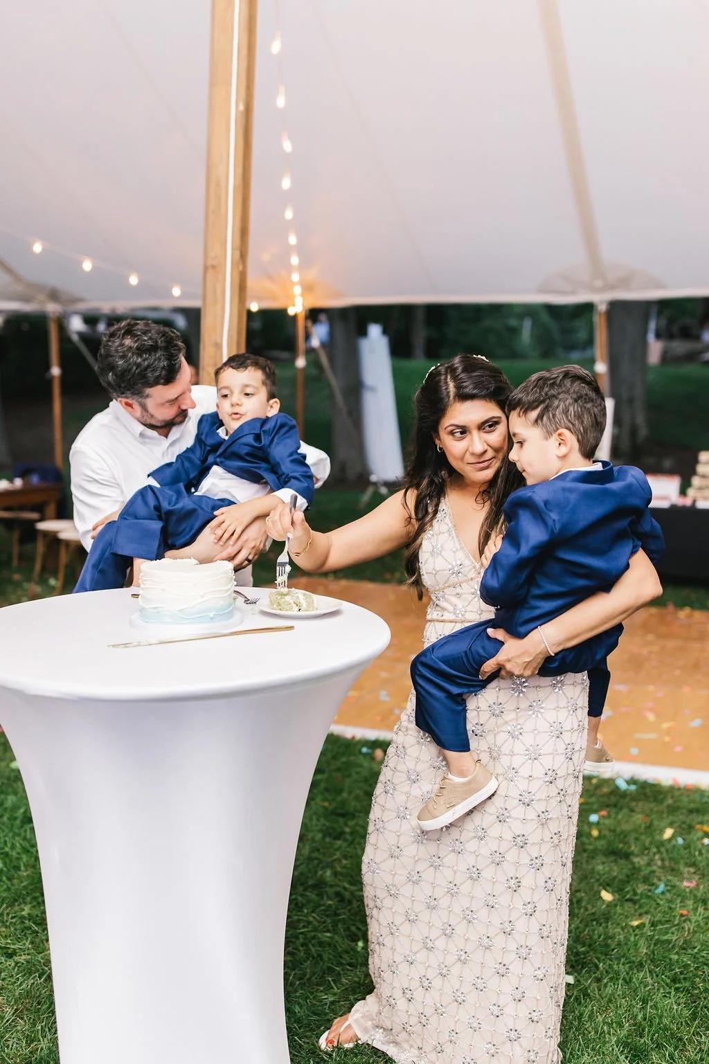 A family celebrating a wedding anniversary under a tent. The woman is holding one of the young boys, while another is being held by a man. A cake is on a high table with a piece on a plate, ready to be served.