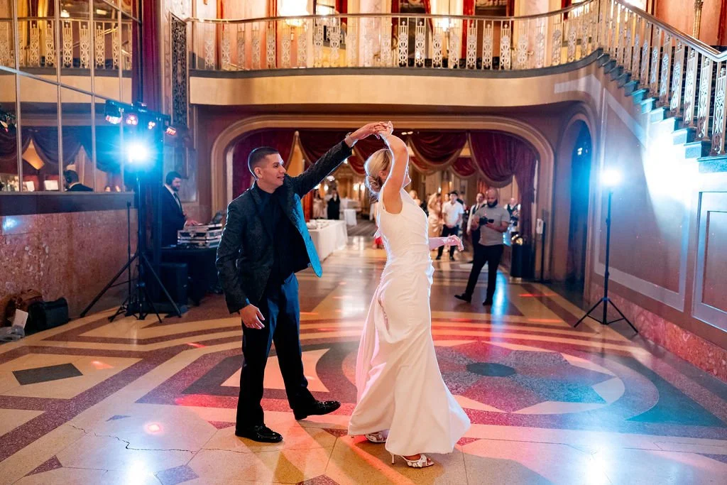 A groom and bride dancing at a wedding reception in a decorated indoor venue with a staircase, chandeliers, and guests in the background.
