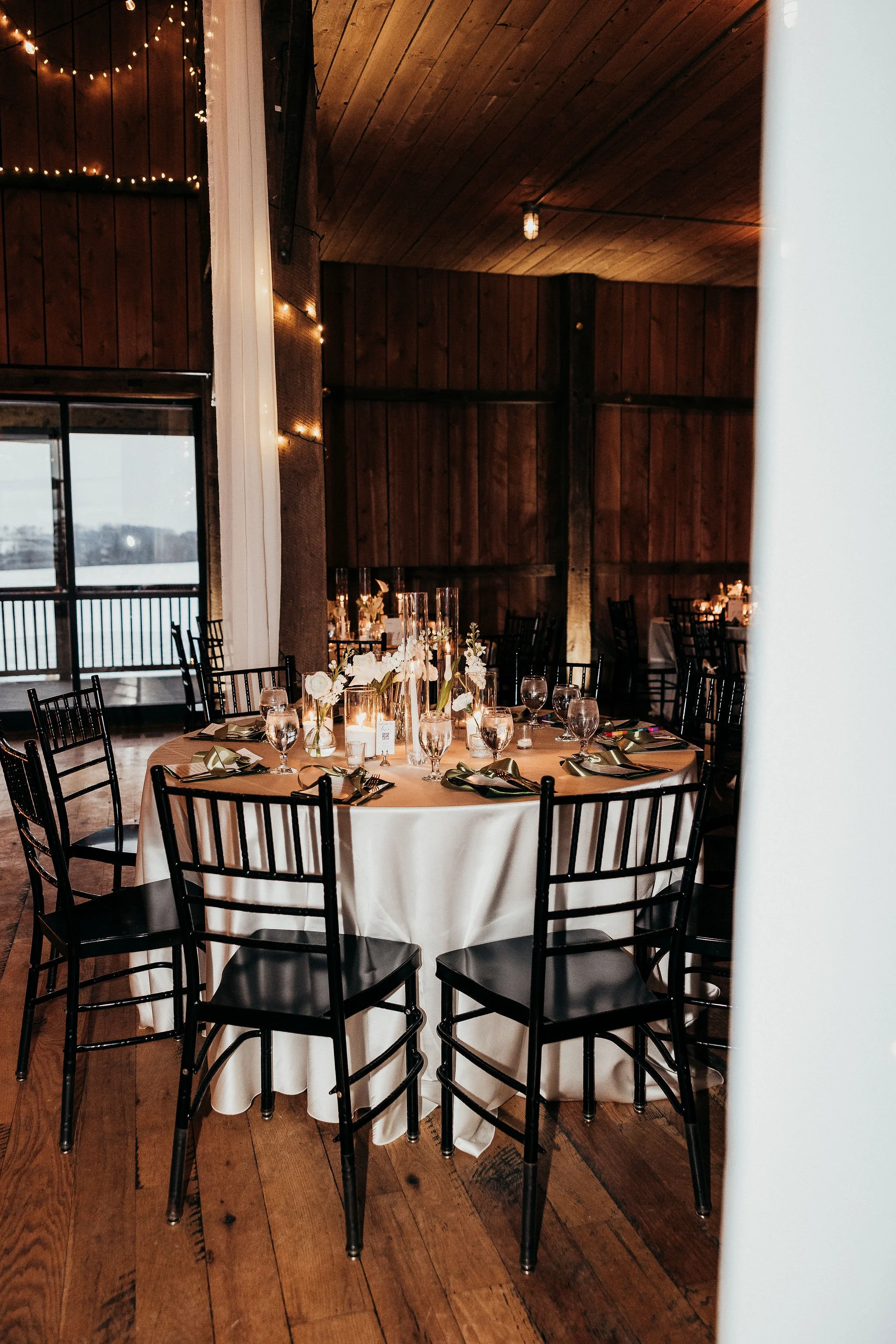 Indoor event space with round table set for a formal dinner, decorated with white flowers in tall vases, candles, and place settings, surrounded by black chairs, with wood-paneled walls and string lights.