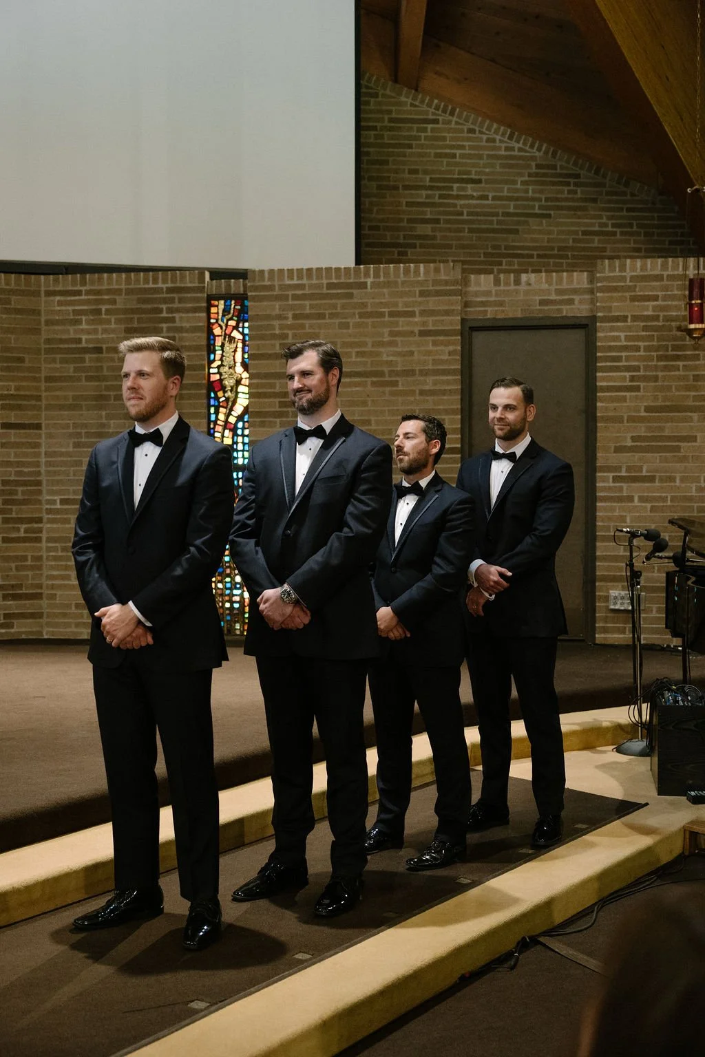 Four men in tuxedos standing in a church or chapel, with a brick wall and stained glass window in the background.