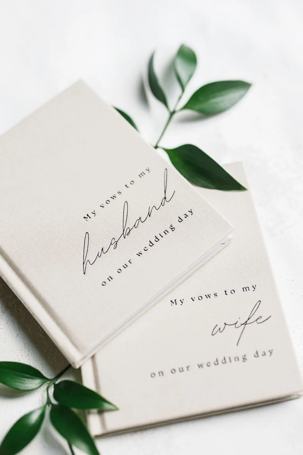 Wedding vow books with green leaves, one with the words 'My vows to my husband on our wedding day' and the other 'My vows to my wife on our wedding day' printed on the covers.