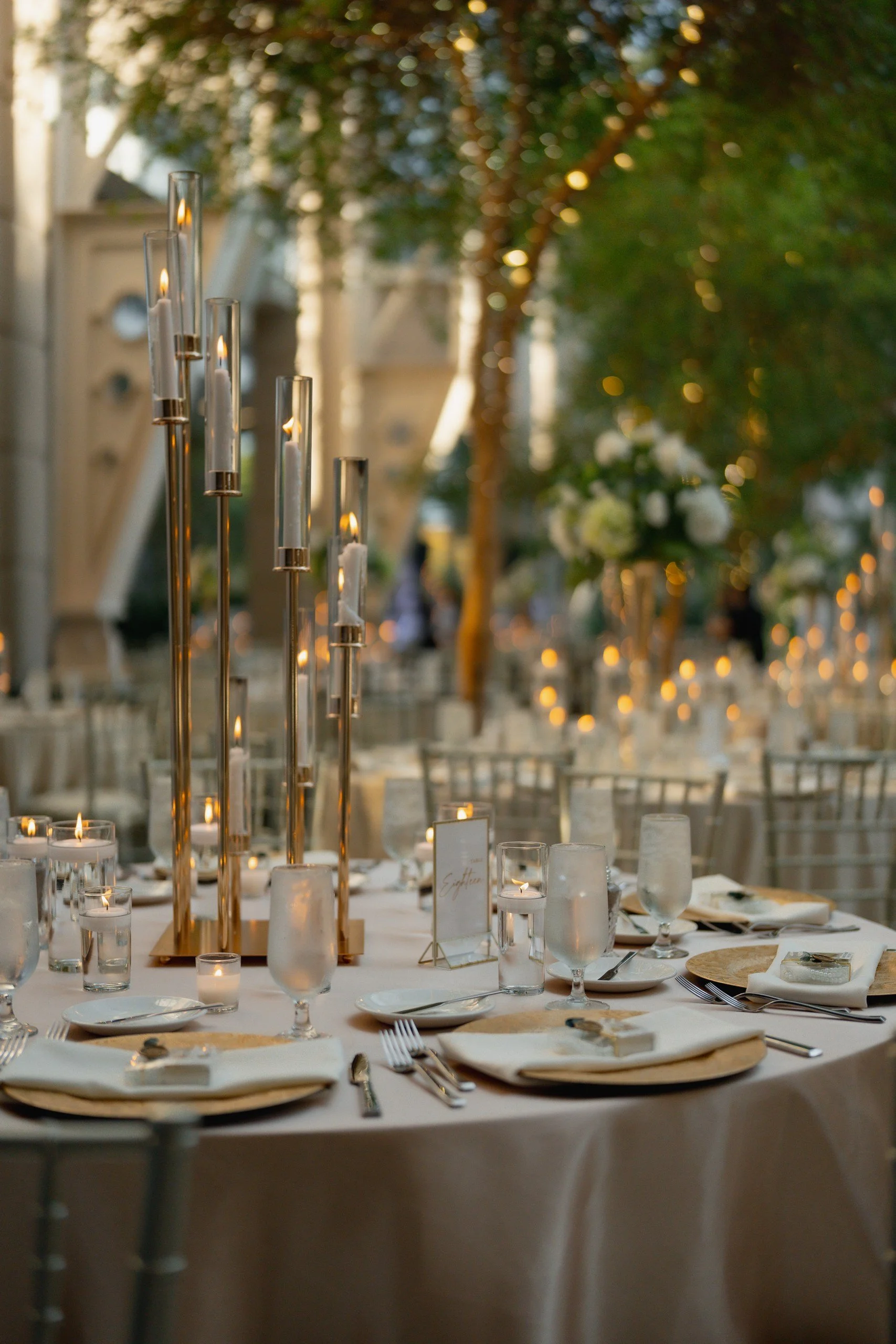 Elegant banquet table set with candles, wine glasses, plates, and cutlery, decorated for a celebration or wedding, with blurred greenery and warm lighting in the background.