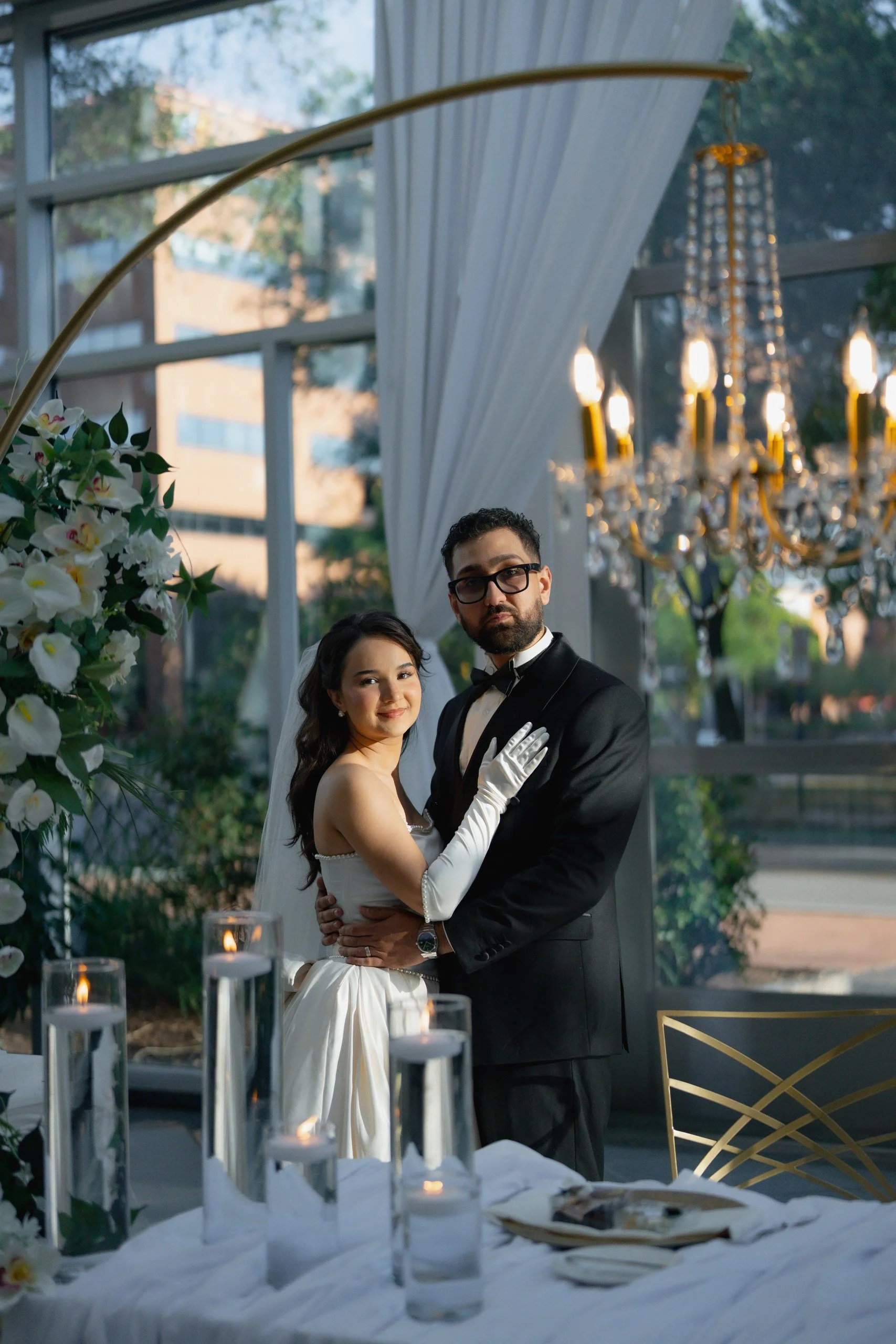 A bride and groom standing together at their wedding reception, with floating candles on a table in the foreground and a chandelier hanging in the background. The scene is set outdoors with large windows and greenery outside.