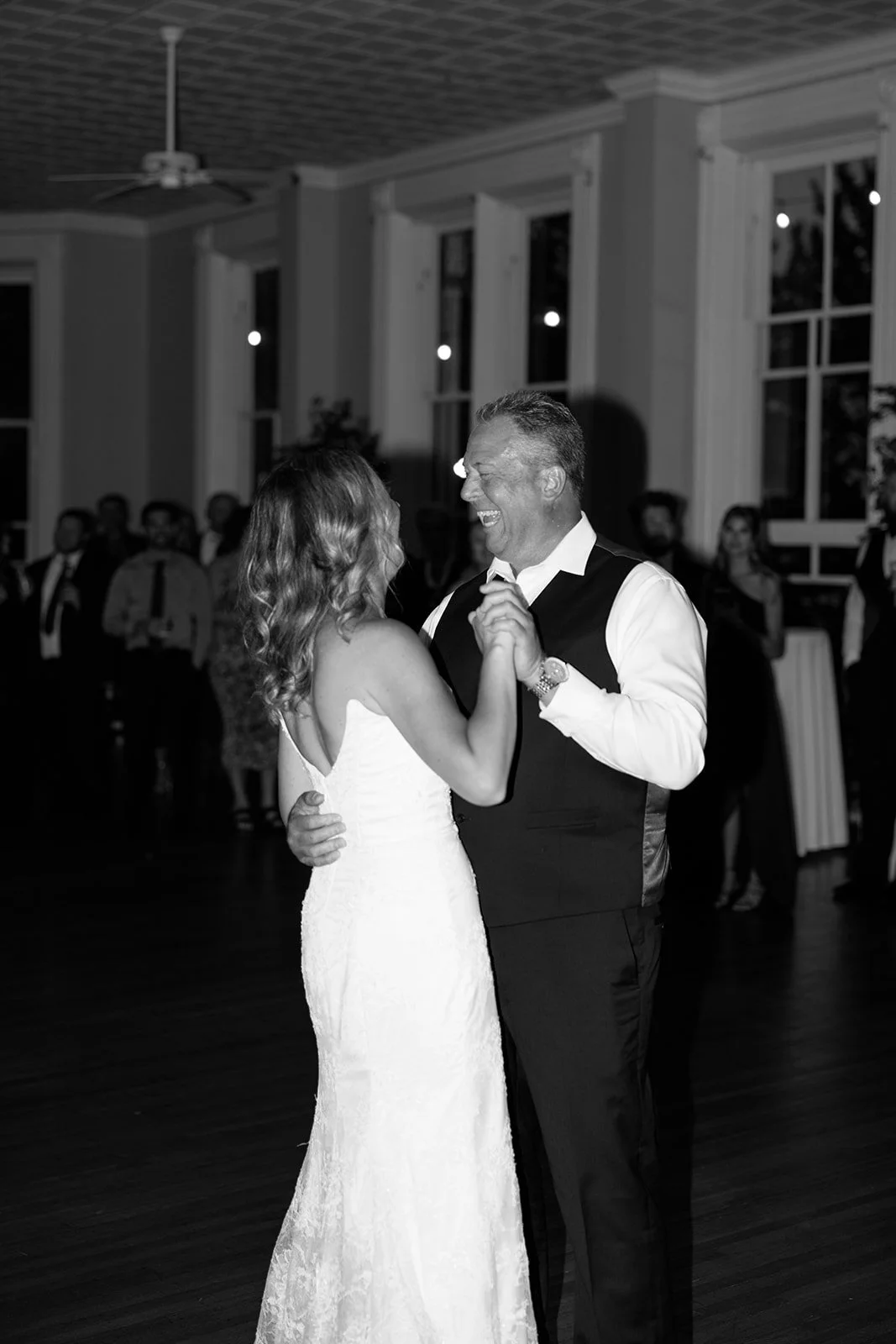 A man and woman dancing, smiling, at a wedding reception, with guests in the background.