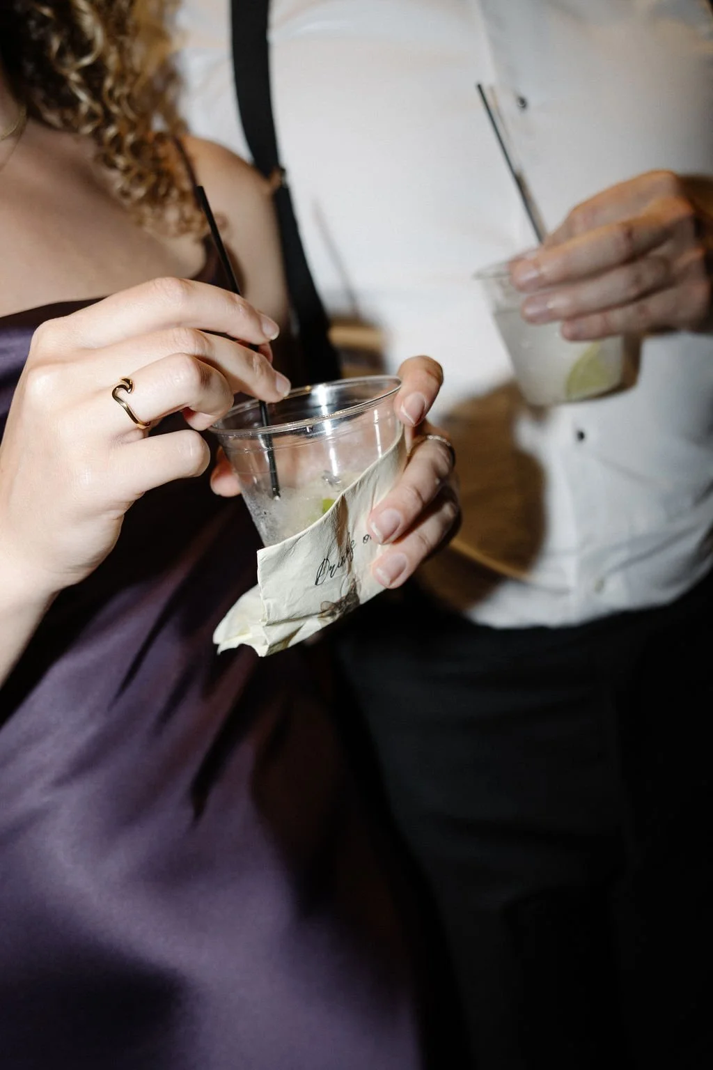 Close-up of two people holding drinks, one with a transparent cup and a lime wedge, the other partially visible, in a social setting.