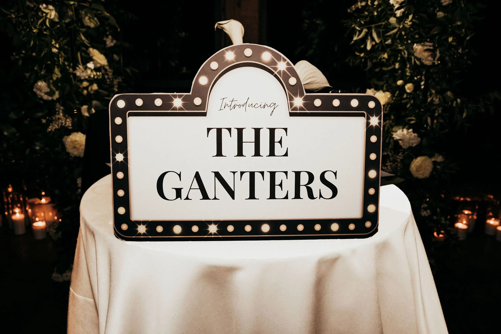 Signboard on a table introducing 'The Ganties' with illuminated border lights, floral background, and candles.
