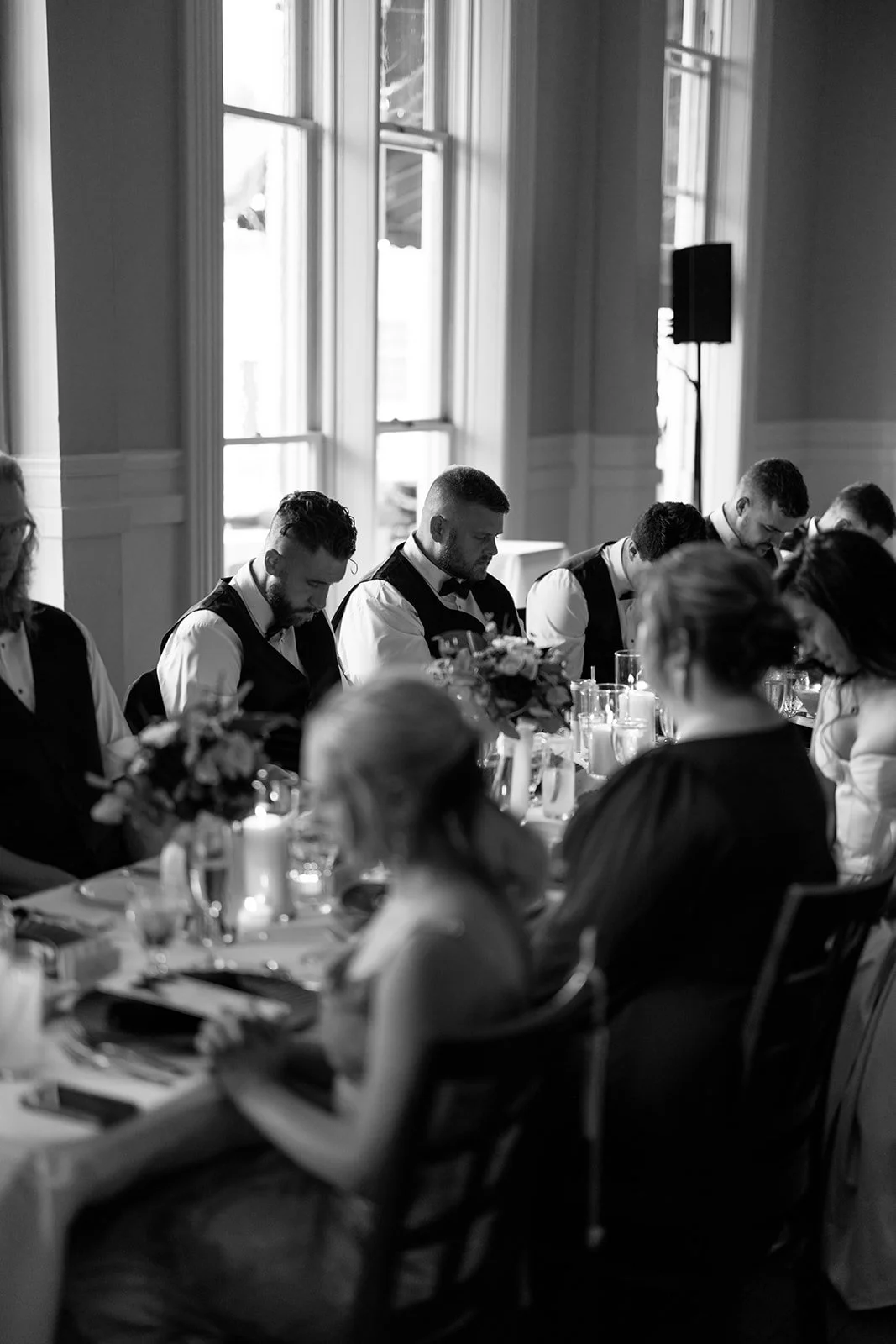 Black and white photo of a group of people dining at a formal event, likely a wedding reception, sitting at a long table with floral centerpieces and candles, in a room with large windows.