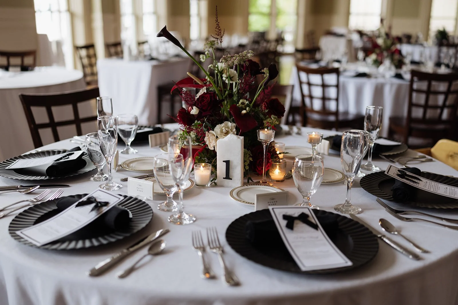 Wedding reception table decorated with black and white plates, black napkins with bows, crystal glassware, candles, and a floral centerpiece with red, white, and green flowers, and a table number.