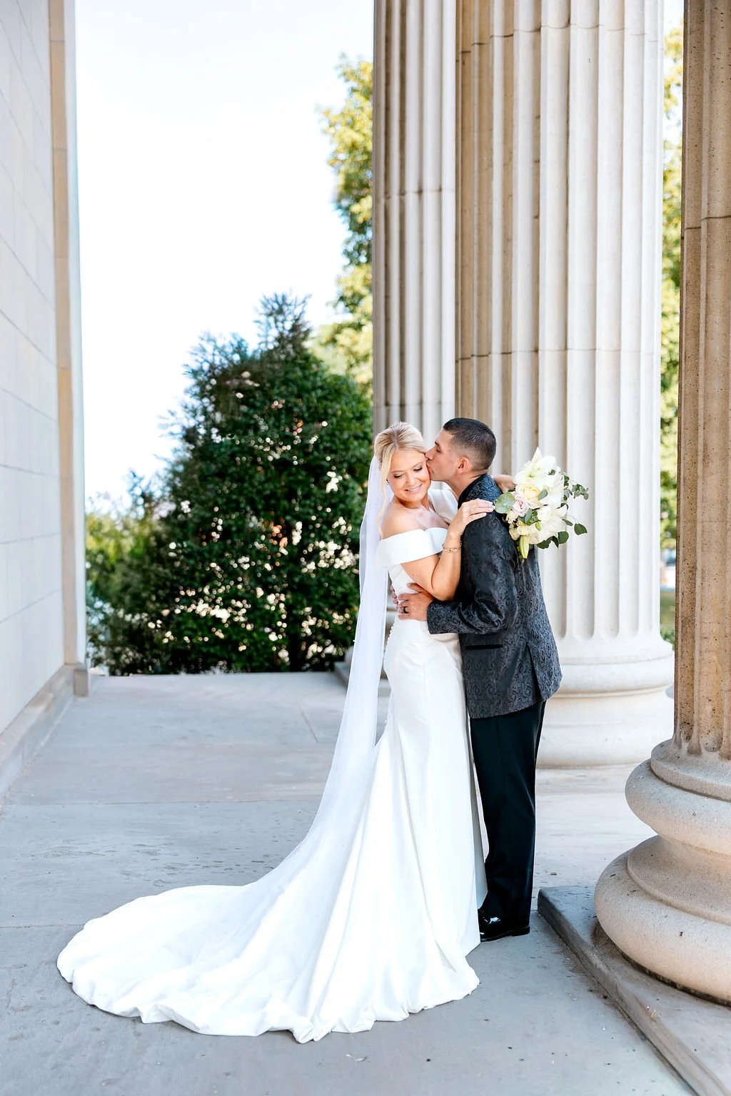 Bride and groom embrace on wedding day, with the groom kissing the bride on the cheek. The bride is holding a bouquet of white and light pink flowers, standing next to large classical columns outside a building.