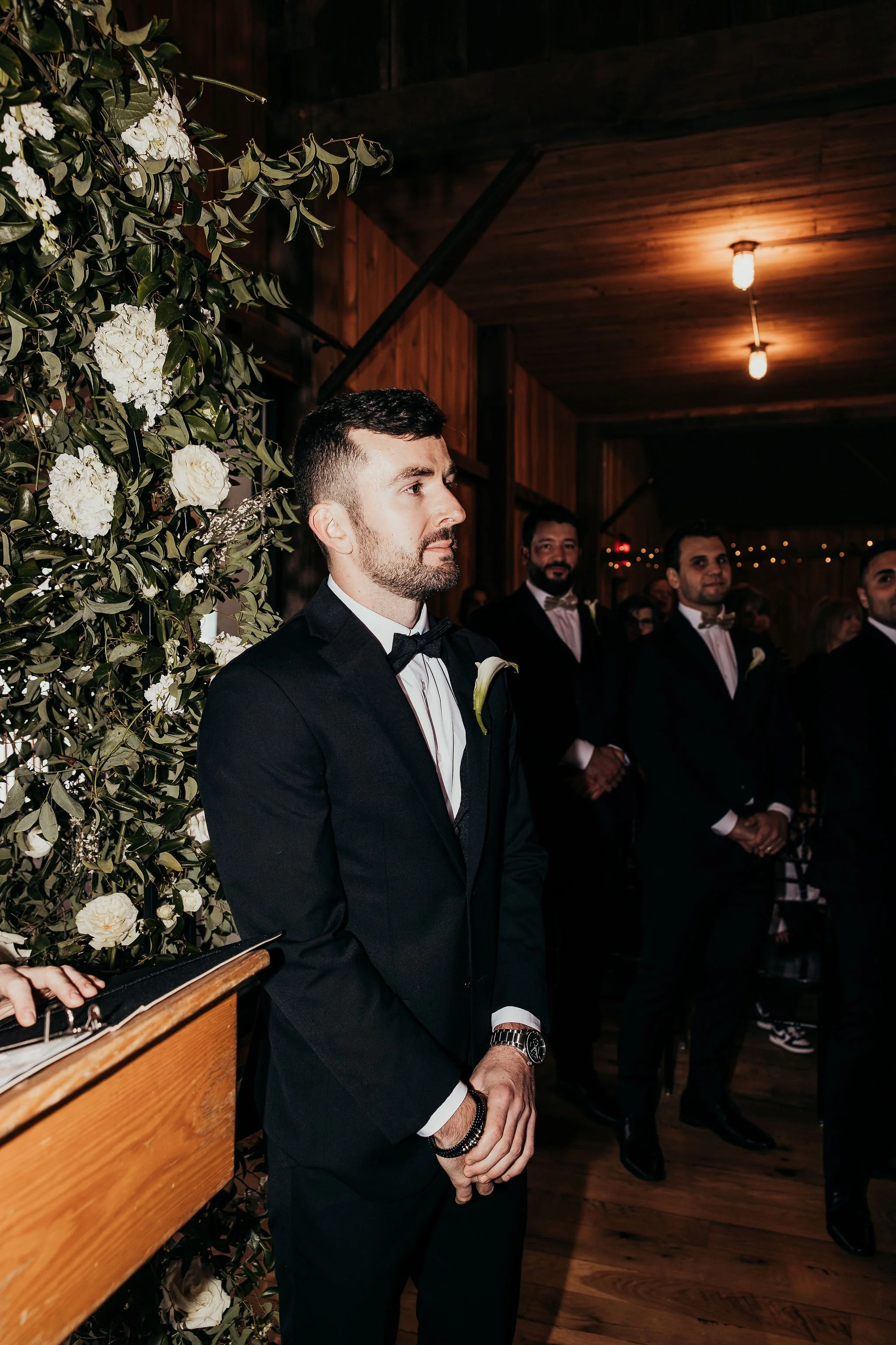 A group of men dressed in tuxedos standing indoors at a formal event, with a decorated floral backdrop on the left and warm lighting on the wooden ceiling.