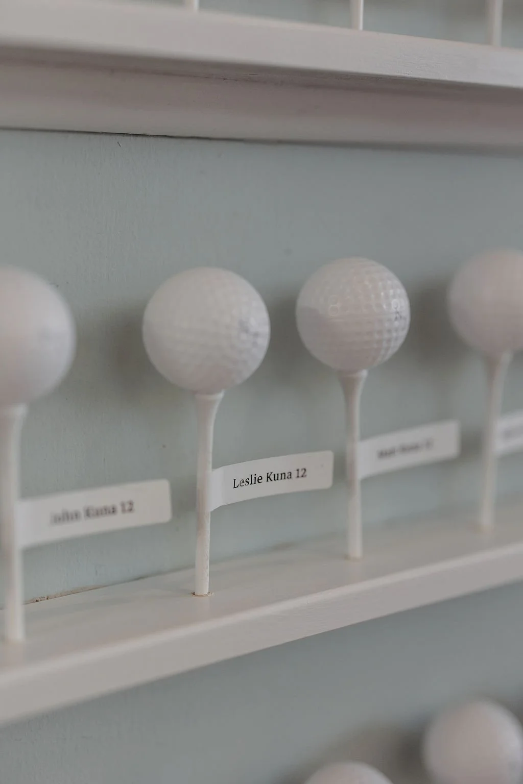 Close-up of white golf tee markers with names and numbers, including Leslie Kuna 12, mounted on a wooden rack.