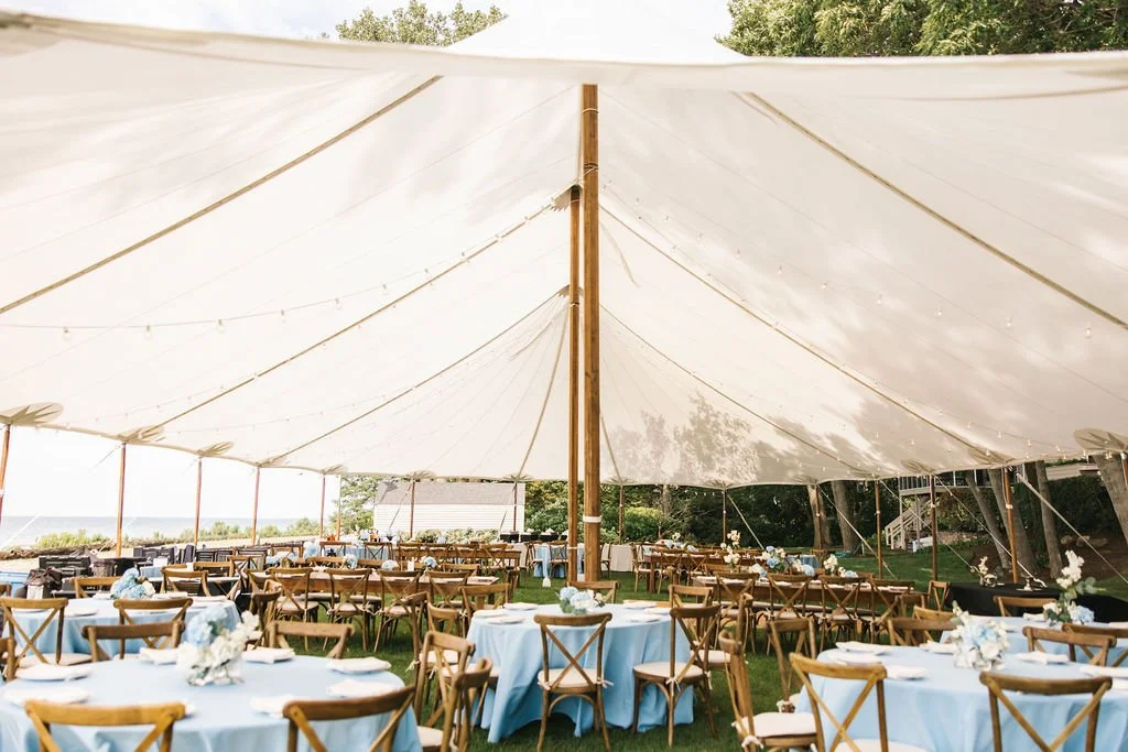 Outdoor event tent with blue-covered tables and wooden chairs, set up for a gathering or celebration, with trees and a grassy area in the background.