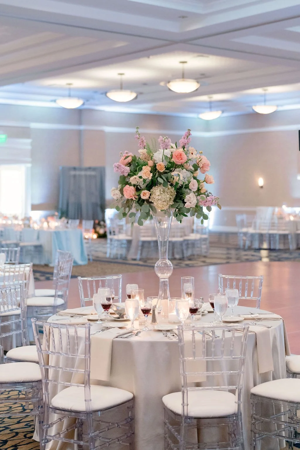 Elegant banquet hall with round tables decorated with white tablecloths, clear chiavari chairs, and tall floral centerpieces featuring pink and white roses and hydrangeas, set for a formal event.