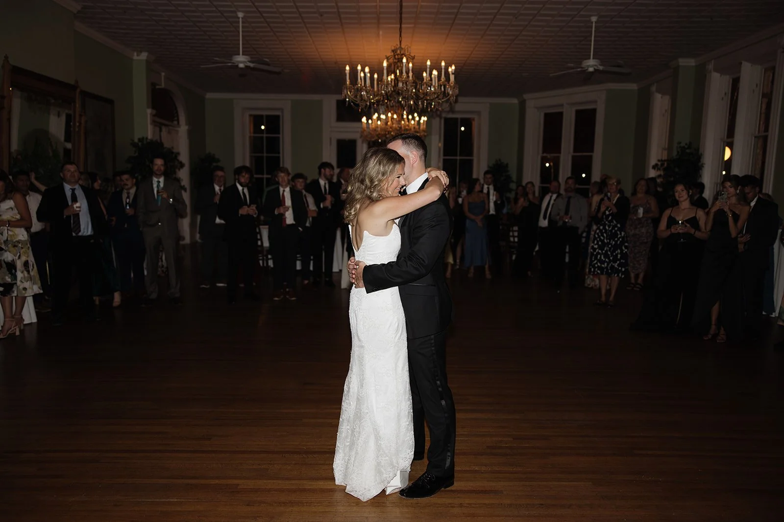 A bride and groom dancing closely at their wedding reception, surrounded by guests watching them in a large, elegant room with chandeliers.