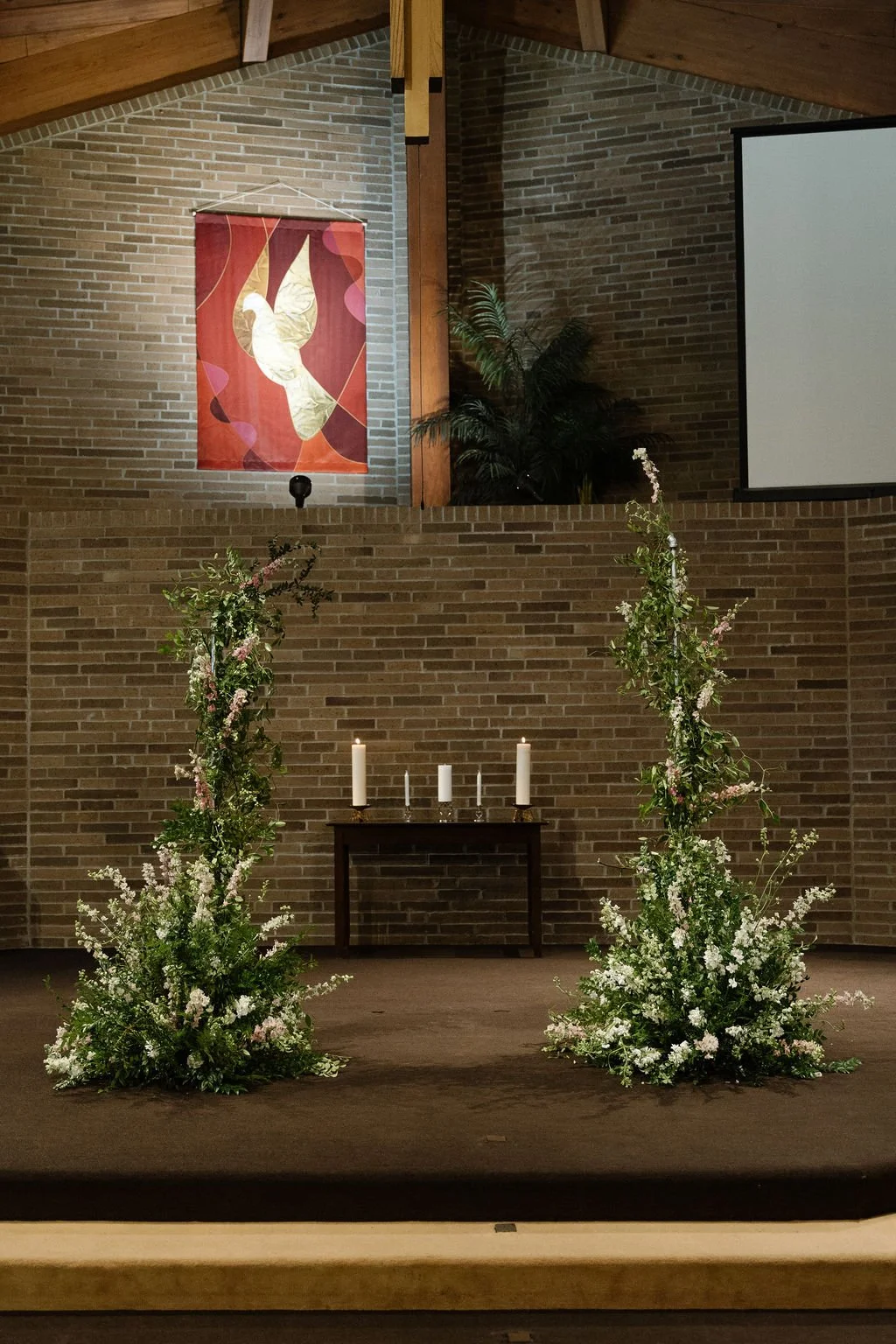 A decorated church altar with two large floral arrangements, three lit candles on a small table, a brick wall backdrop, a hanging abstract art piece, and a projection screen.