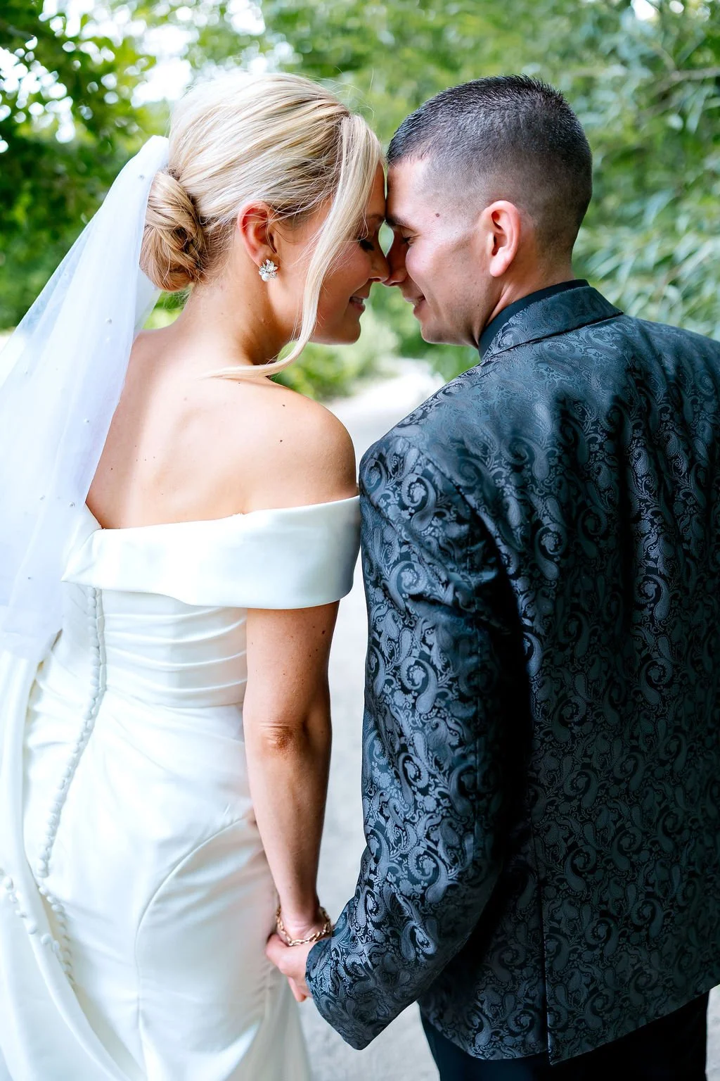 Bride and groom standing close, touching foreheads, holding hands, outdoor setting with greenery, wedding attire.