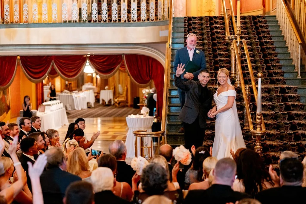 Wedding ceremony with bride and groom at the staircase, officiant standing behind them, guests watching and taking pictures in an elegant ballroom with red curtains and a grand staircase.
