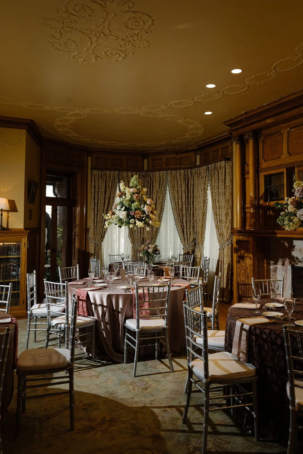 Elegant dining room with round table set for a formal event, adorned with a large floral centerpiece, surrounded by silver chairs, with gold and patterned curtains and wooden paneling in the background.
