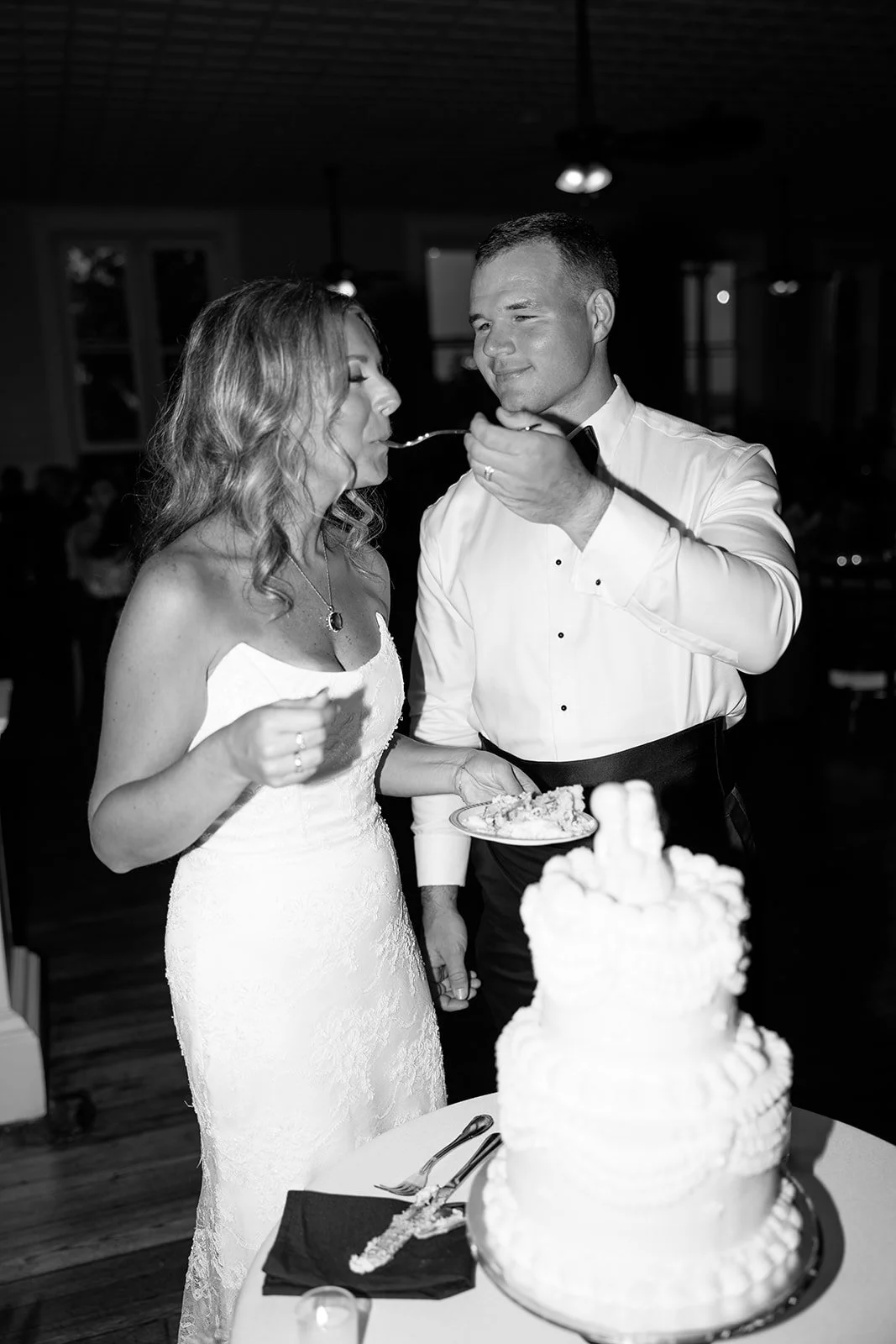 A black and white photo of a couple at their wedding reception, the woman in a white strapless wedding dress with wavy hair, and the man in a white dress shirt with a black bow tie. They are standing close, with the man feeding the woman cake, and a 