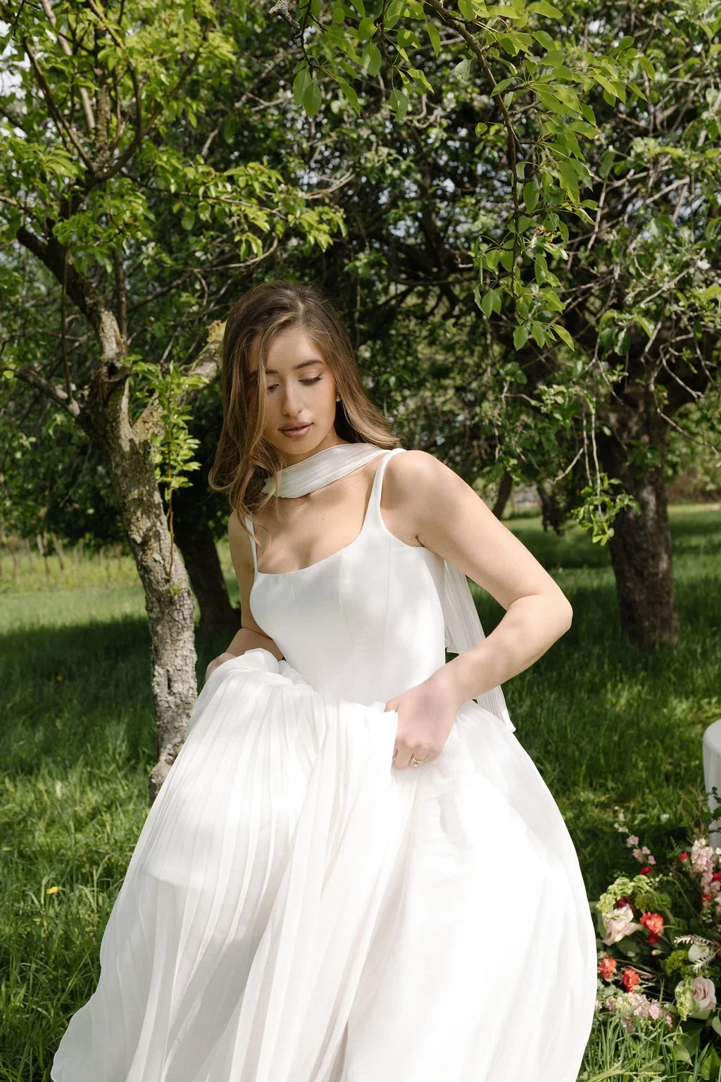 A young woman in a white dress standing outdoors near a tree, with green grass and flowers around her.