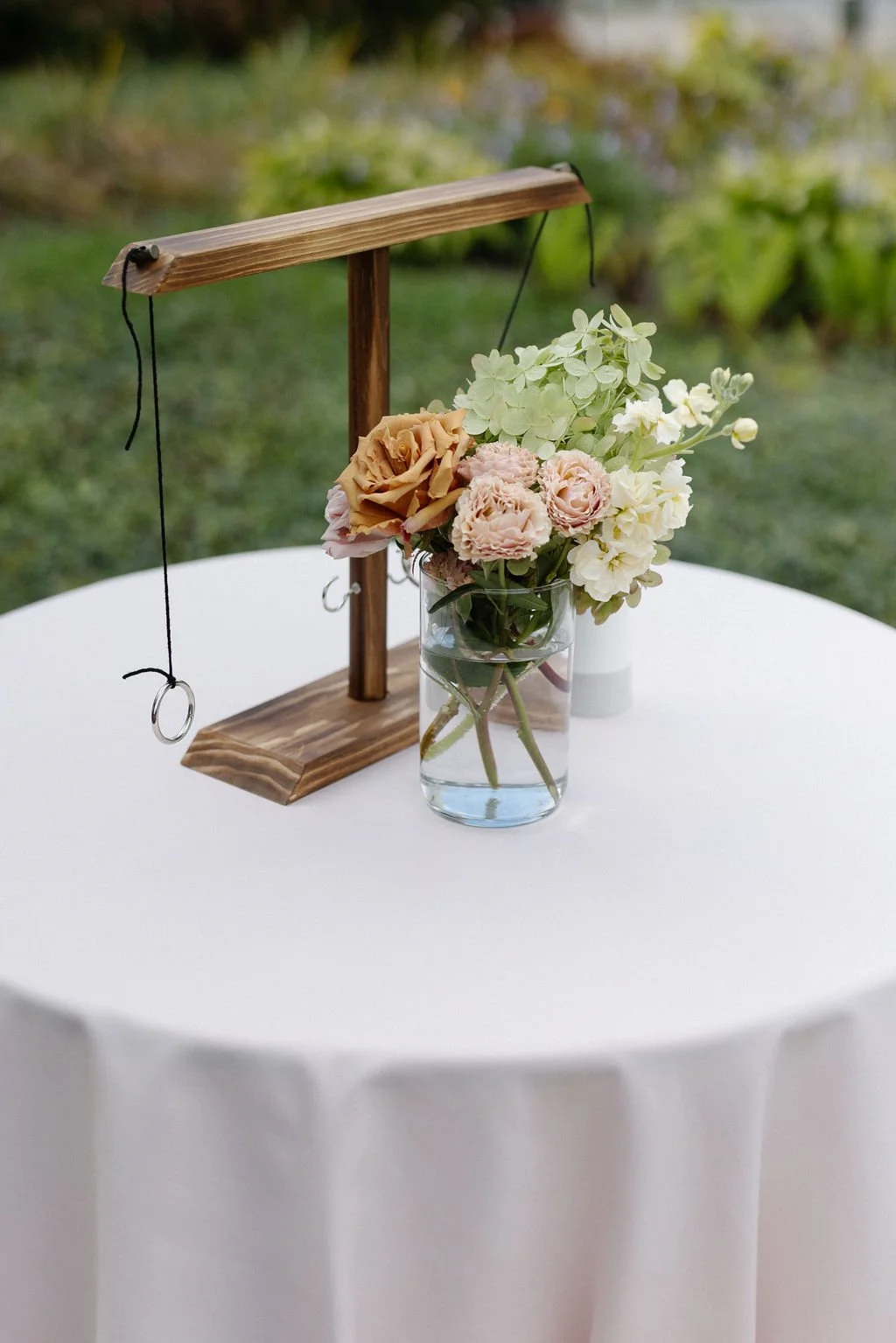 A table with a floral arrangement in a glass vase and a jewelry display stand, outdoors with greenery in the background.