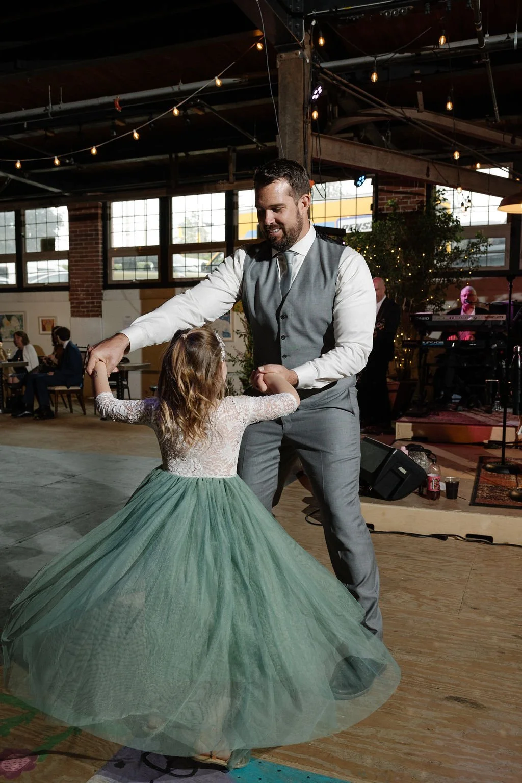 A man and a young girl are dancing together at an indoor event. The man is dressed in a gray vest and pants, and the girl is wearing a long green tulle dress with a lace top. The background shows other people sitting at tables and a band performing o