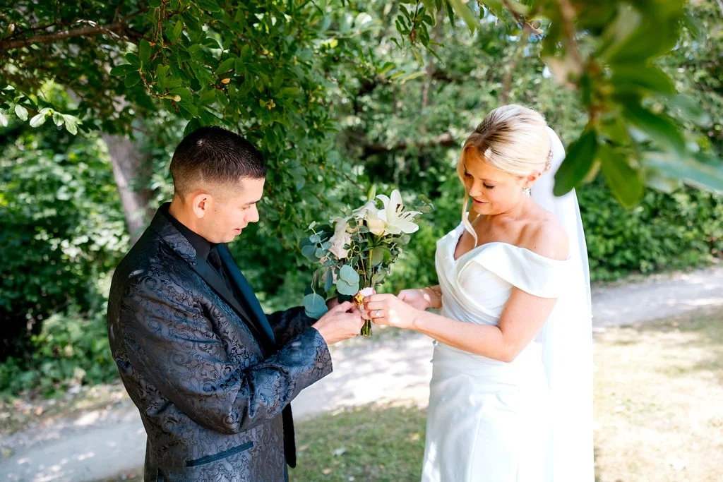A bride in a white wedding dress and a groom in a dark suit exchange rings outdoors under trees.