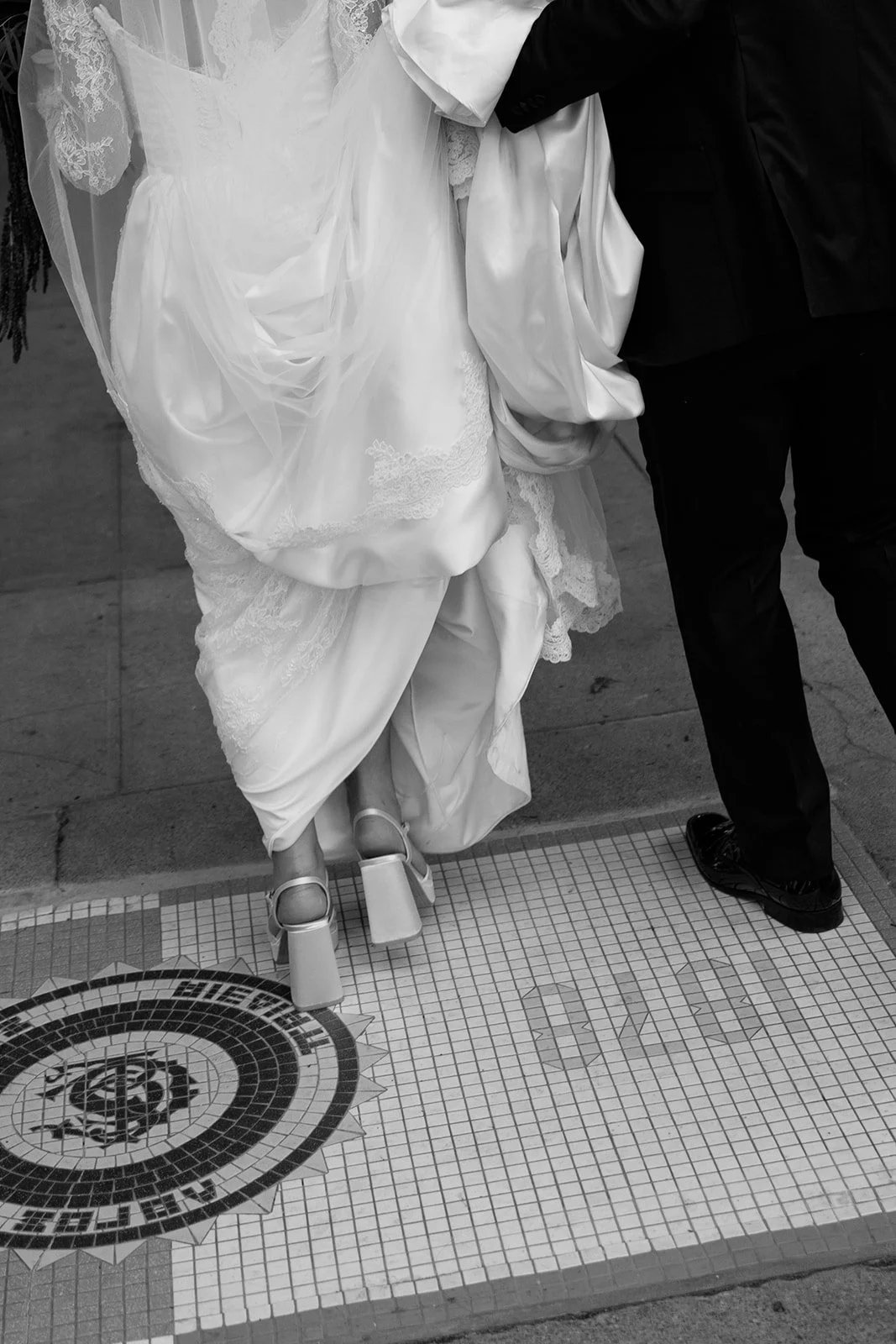Close-up of a bride in a wedding gown and a groom in a suit, walking together on a tiled sidewalk.