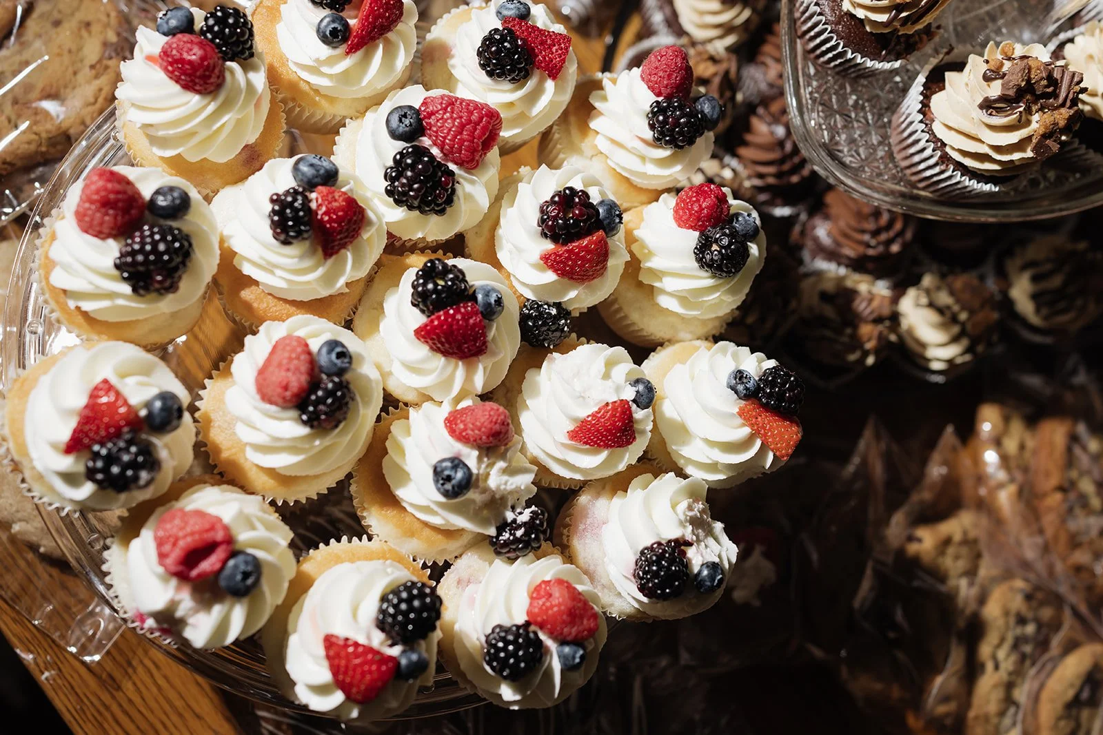 A tray of mini vanilla cupcakes topped with white frosting and mixed berries, including raspberries, blackberries, and blueberries, arranged on a glass platter.