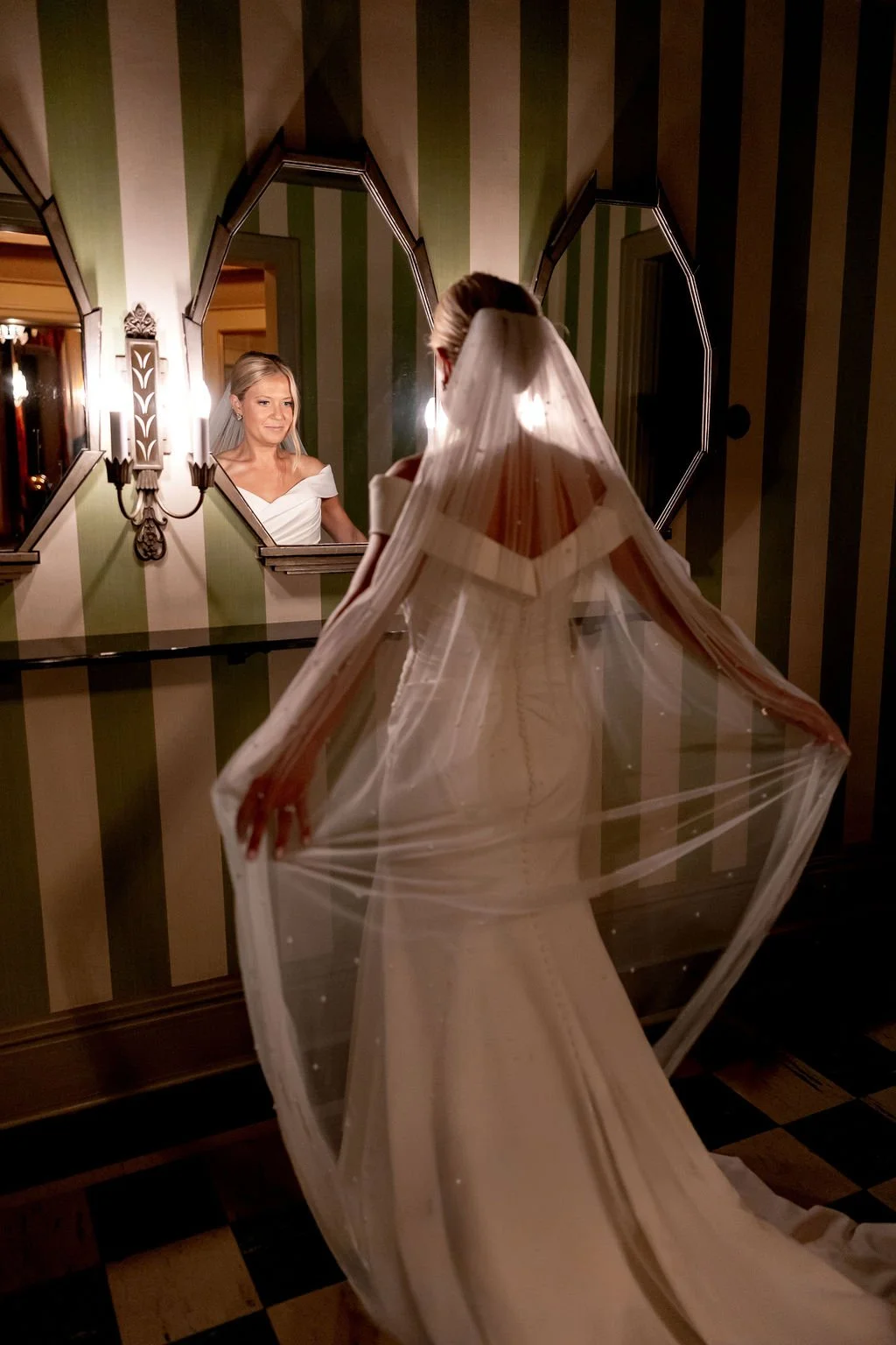 A bride in a wedding dress with a veil looking at herself in a mirror in a decorated room.