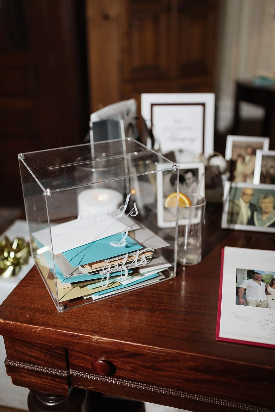 A clear acrylic box filled with colorful envelopes sits on a wooden table, with a glass of water containing a lemon slice nearby. In the background, framed photographs and a handwritten note are visible.