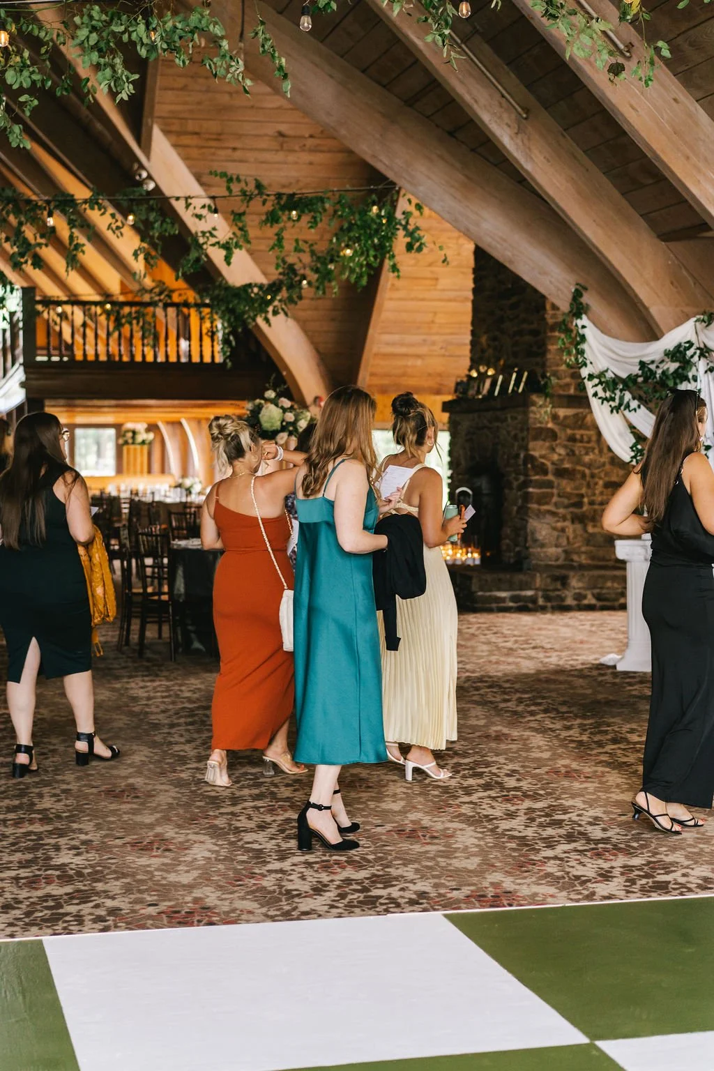 Group of women in formal attire standing inside a rustic wooden venue with a high arched ceiling, greenery, and string lights.
