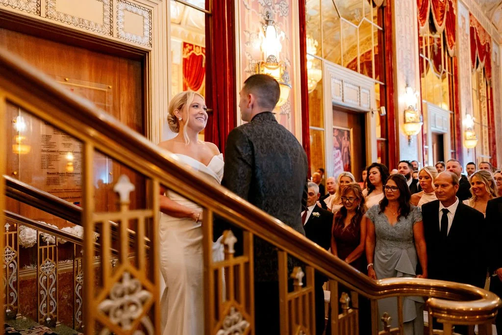 A bride and groom exchanging vows at their wedding ceremony inside a grand, ornate hall with red curtains and gold details, as guests look on.