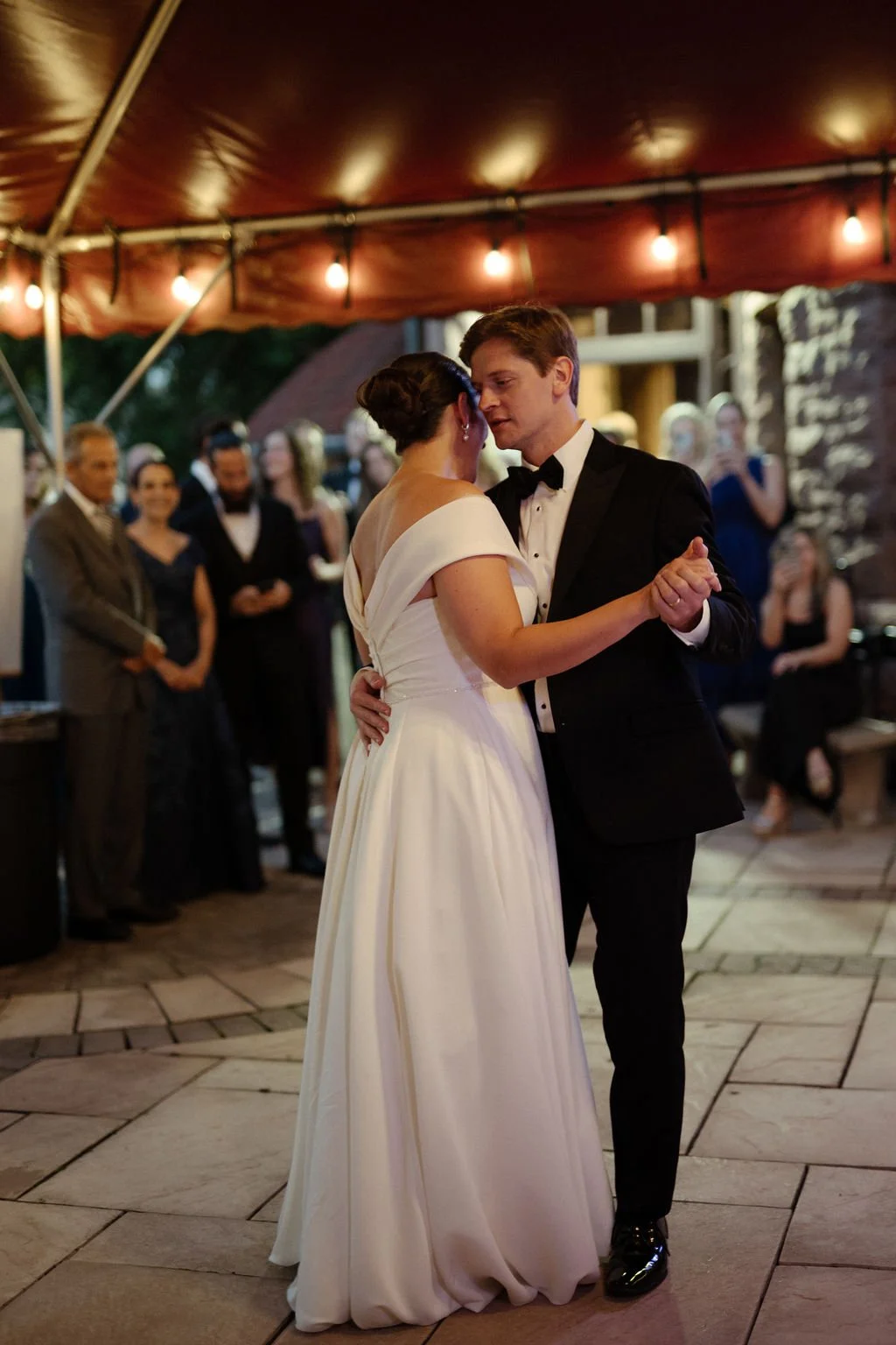A bride and groom have their first dance at a wedding reception, surrounded by guests under a canopy with string lights.