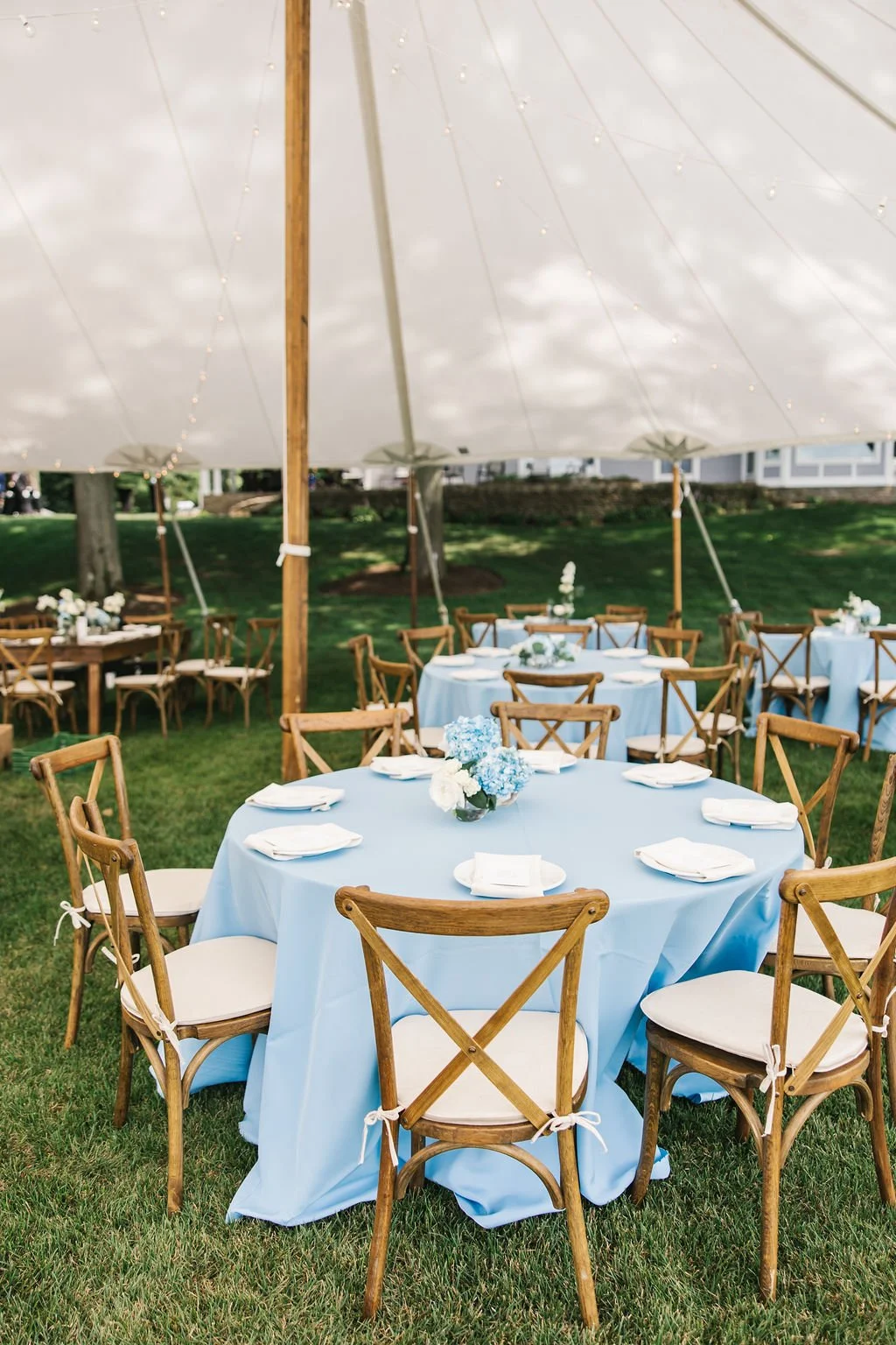 Outdoor event setup with round tables covered in light blue tablecloths, each with white napkins and floral centerpieces, under a large white canopy tent with wooden support beams, on a grassy area.