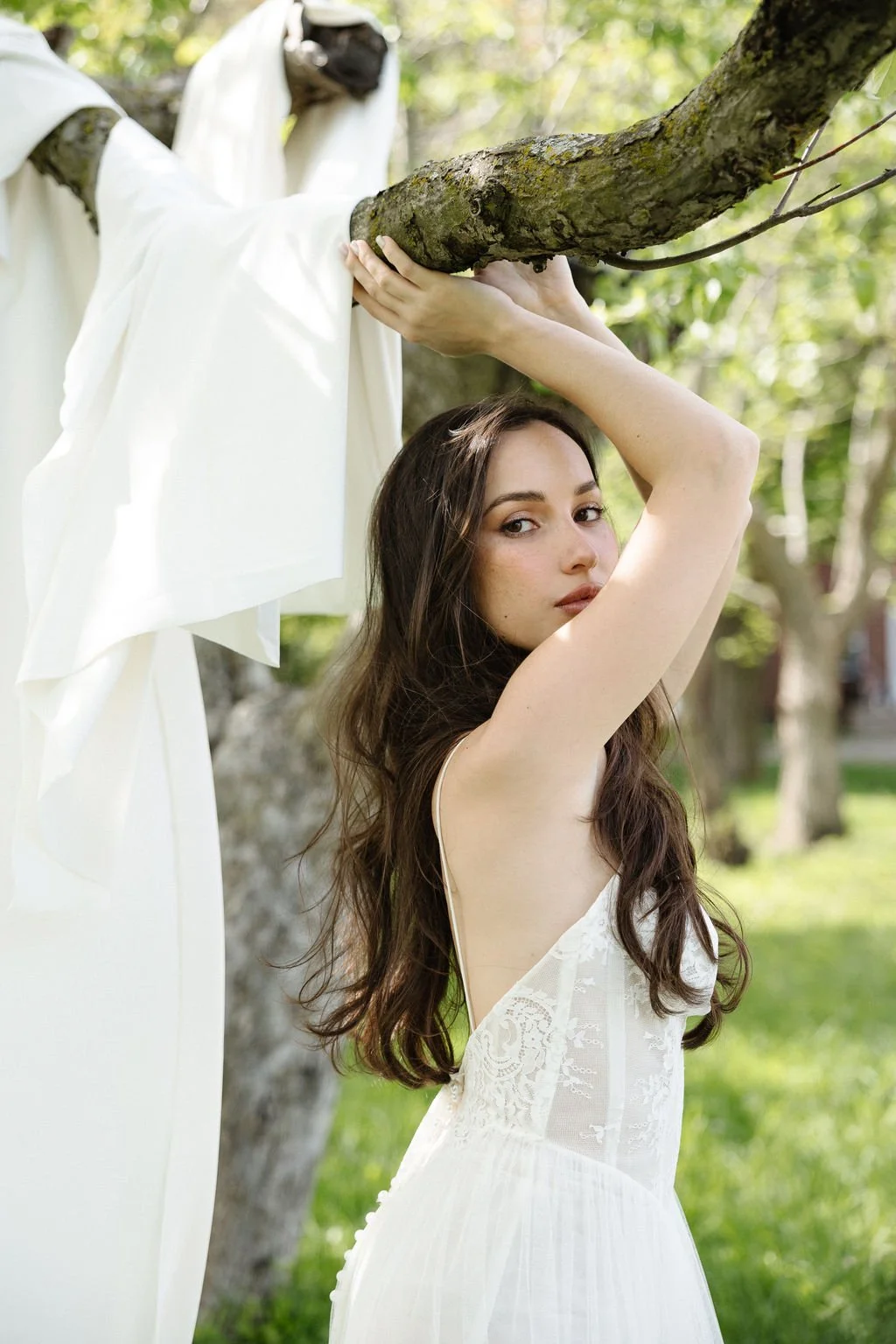 A woman in a white lace dress leaning against a tree with a white cloth draped over a branch, outdoors during daylight.