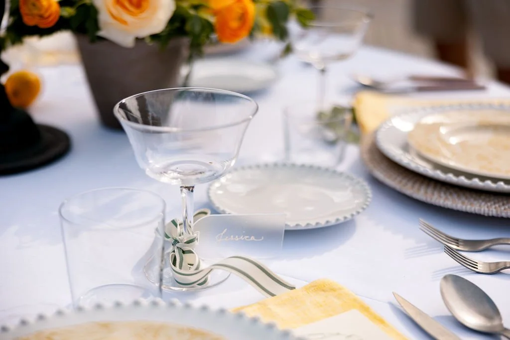 Elegant table setting with glassware, white plates with scalloped edges, silverware, and a floral centerpiece with yellow and white flowers.