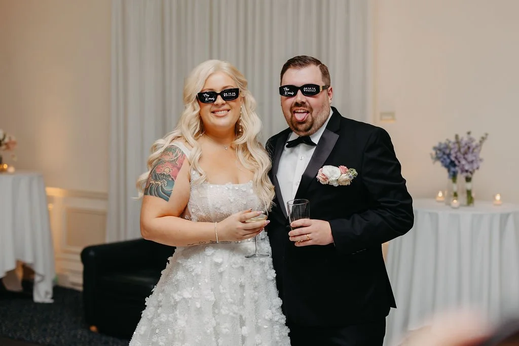 Couple at wedding reception wearing blackout glasses, with one woman in a white wedding dress and one man in a black tuxedo, smiling and posing for the camera.