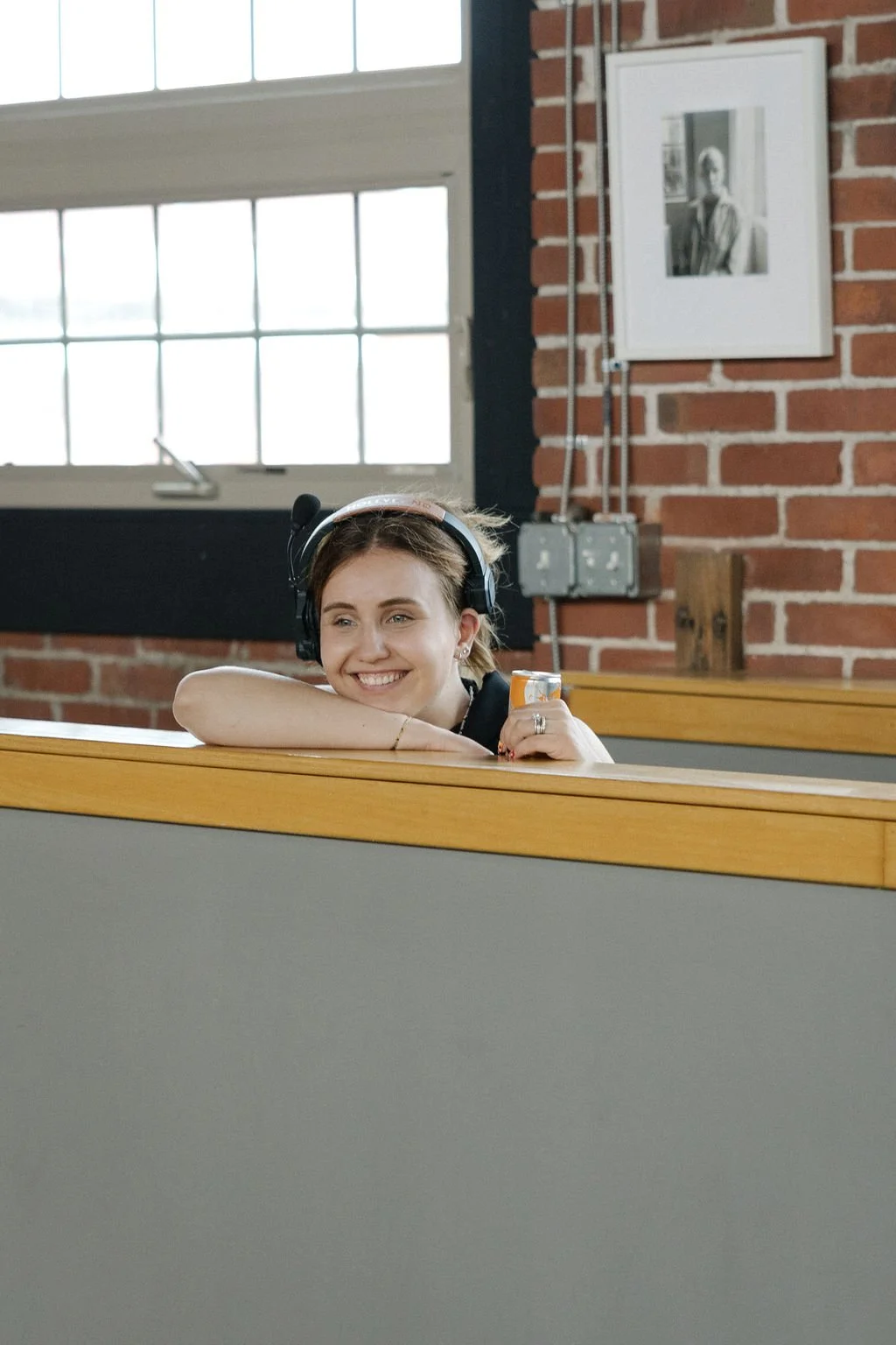 A young woman laughing and smiling with her chin resting on her arms, wearing headphones and holding an orange soda can in a room with brick walls and large windows