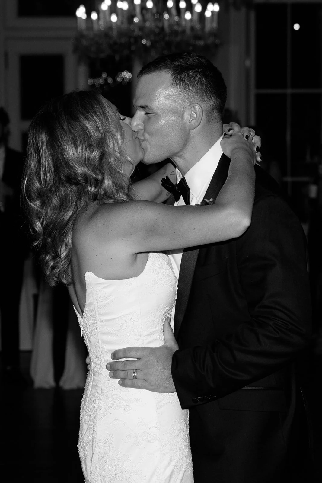 A Black and white photo of a bride and groom kissing on the dance floor at their wedding reception, with the bride wearing a lace wedding dress and the groom in a tuxedo.