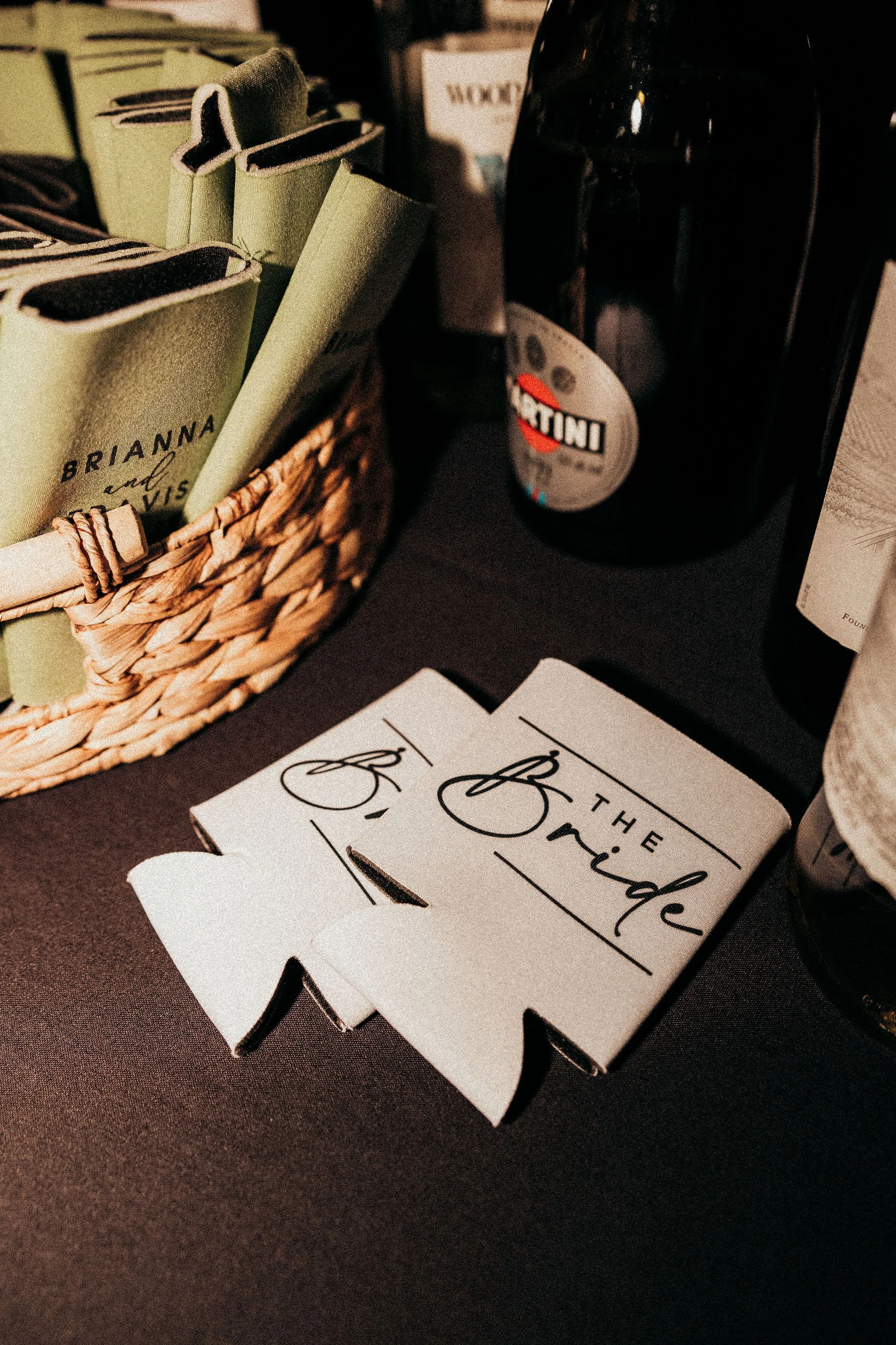 A basket with folded green cloth napkins and a black bottle, with a white card that reads "The Bachelorette" on a dark surface.