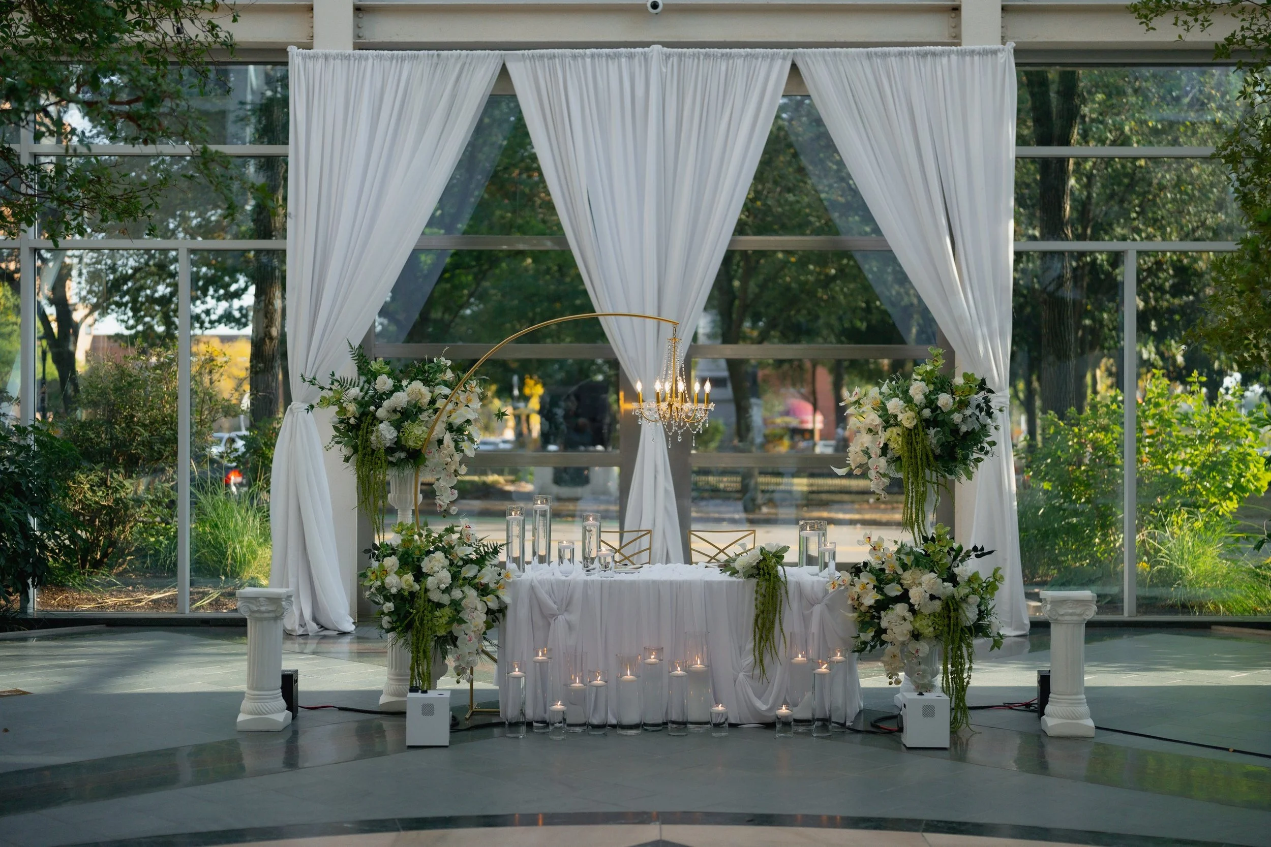 Elegant wedding altar setup with white drapes, floral arrangements, and a chandelier, set outdoors with trees in the background.