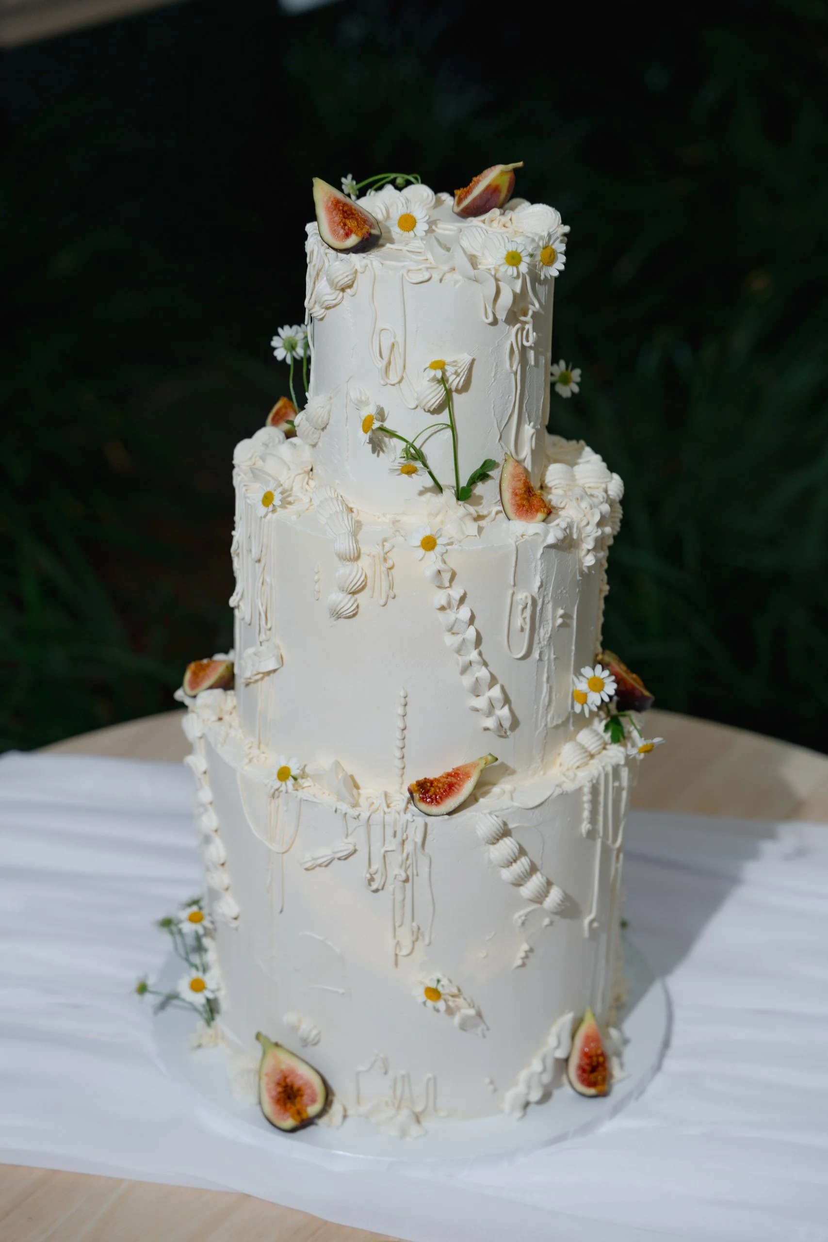 Two-tier white wedding cake decorated with fresh figs and small white daisies, sitting on a white cake board with a white tablecloth underneath.