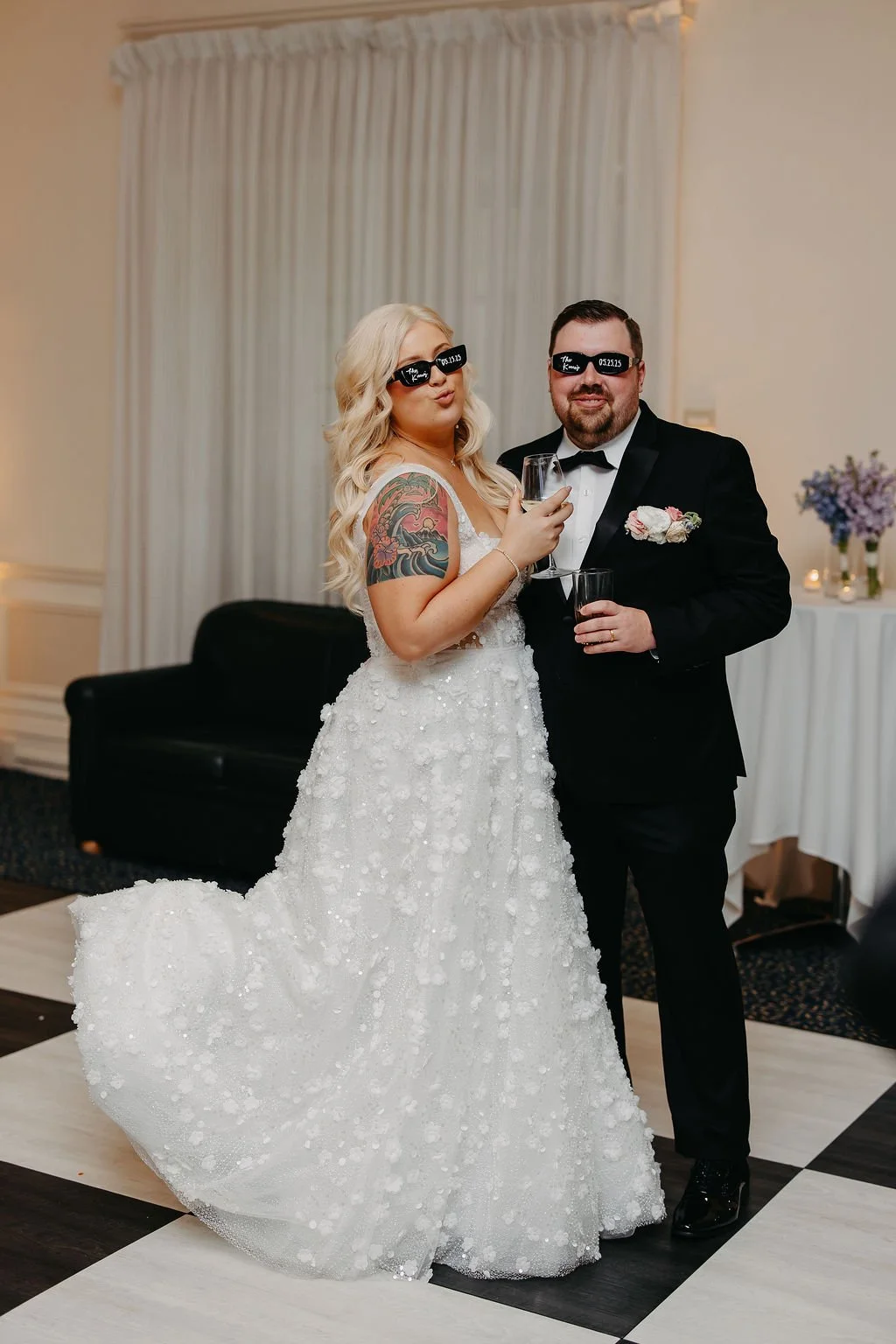 Bride and groom at wedding reception wearing sunglasses, holding glasses of champagne, standing on black and white checkered dance floor, with cream-colored curtains and decorated table in the background.