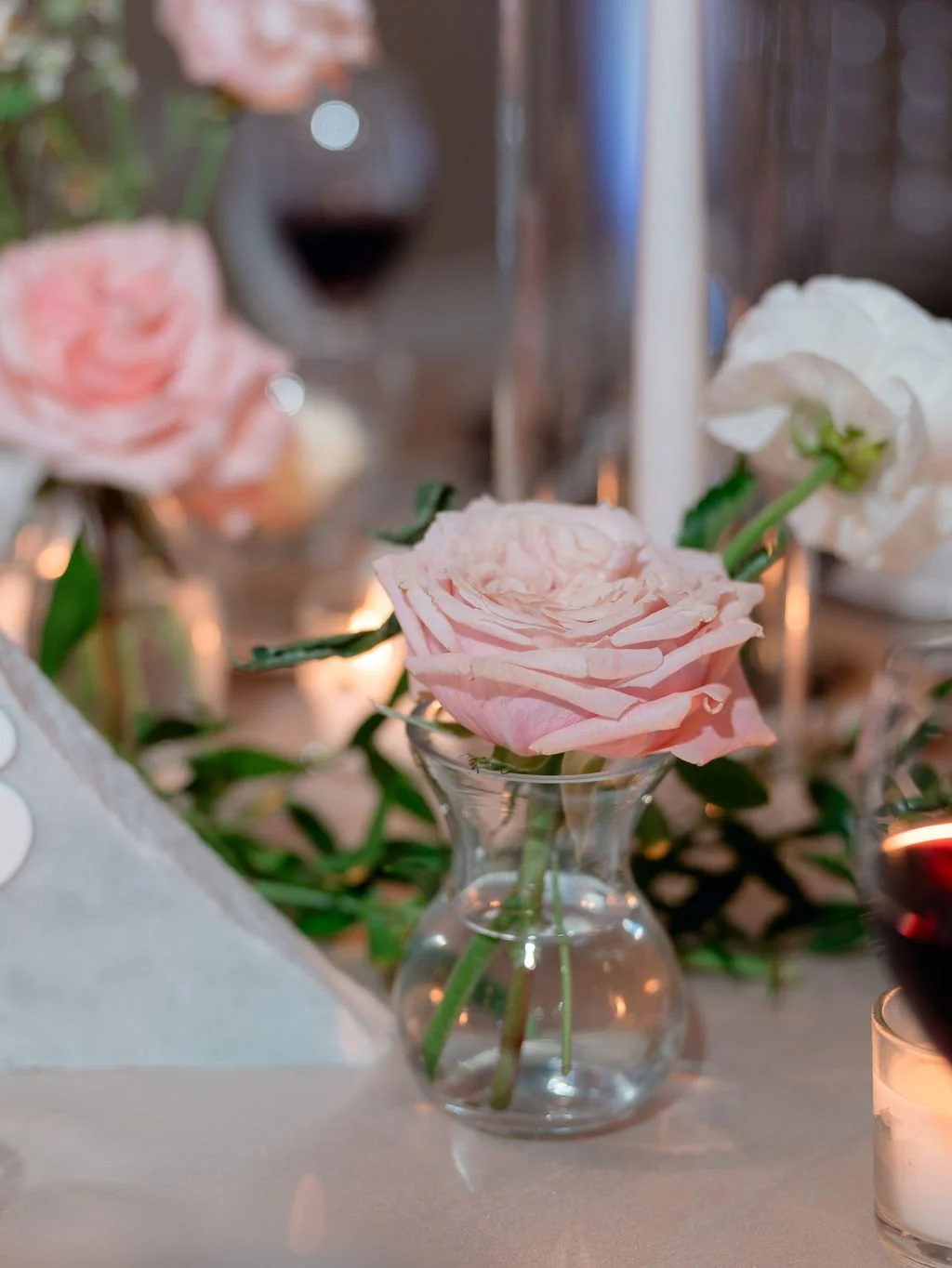 Close-up of pink and white roses in small glass vases on a table with a blurred background of a wine glass and a lit candle.